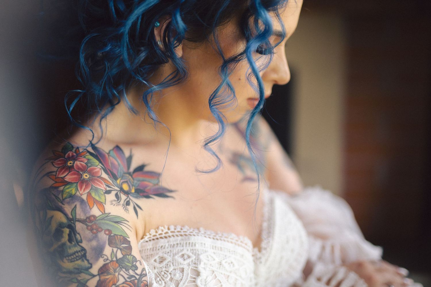 A bride in a white lace dress with blue curled hair and colorful floral tattoos on her shoulder.