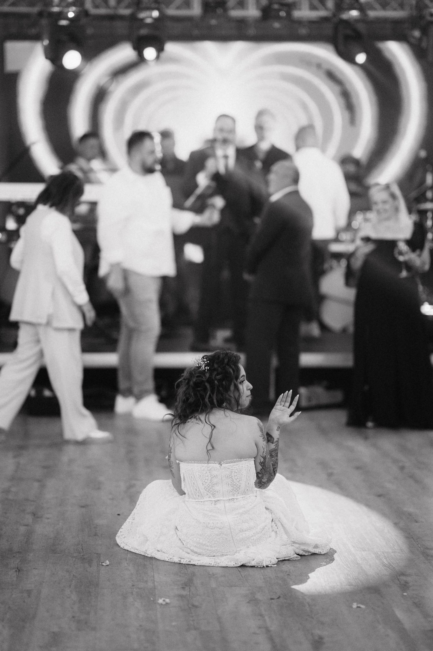 A bride in a white dress sits on a wooden dance floor watching others dance at a reception with circular lights in background.