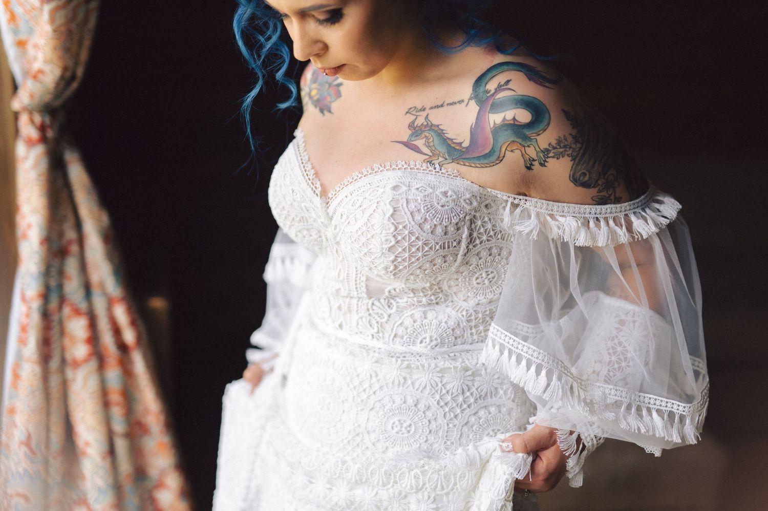 A close-up view of a white lace wedding dress with sheer sleeves and visible shoulder tattoo artwork.