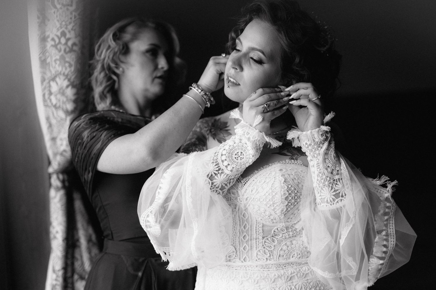 Black and white photo shows a bride in lace white dress getting help with jewelry before her wedding ceremony.