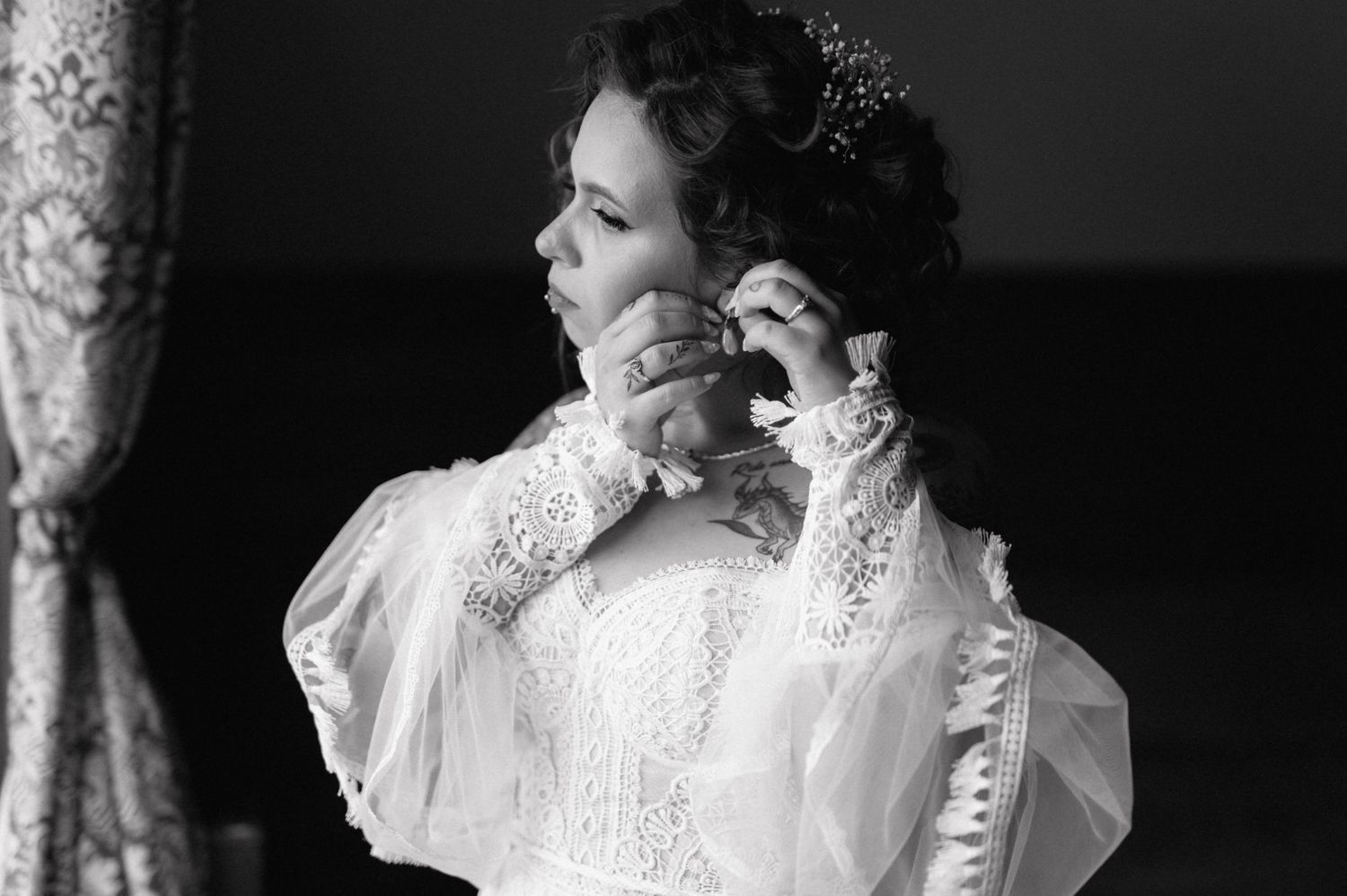 Black and white photograph of a bride in a vintage lace wedding dress fastening an earring during preparations.