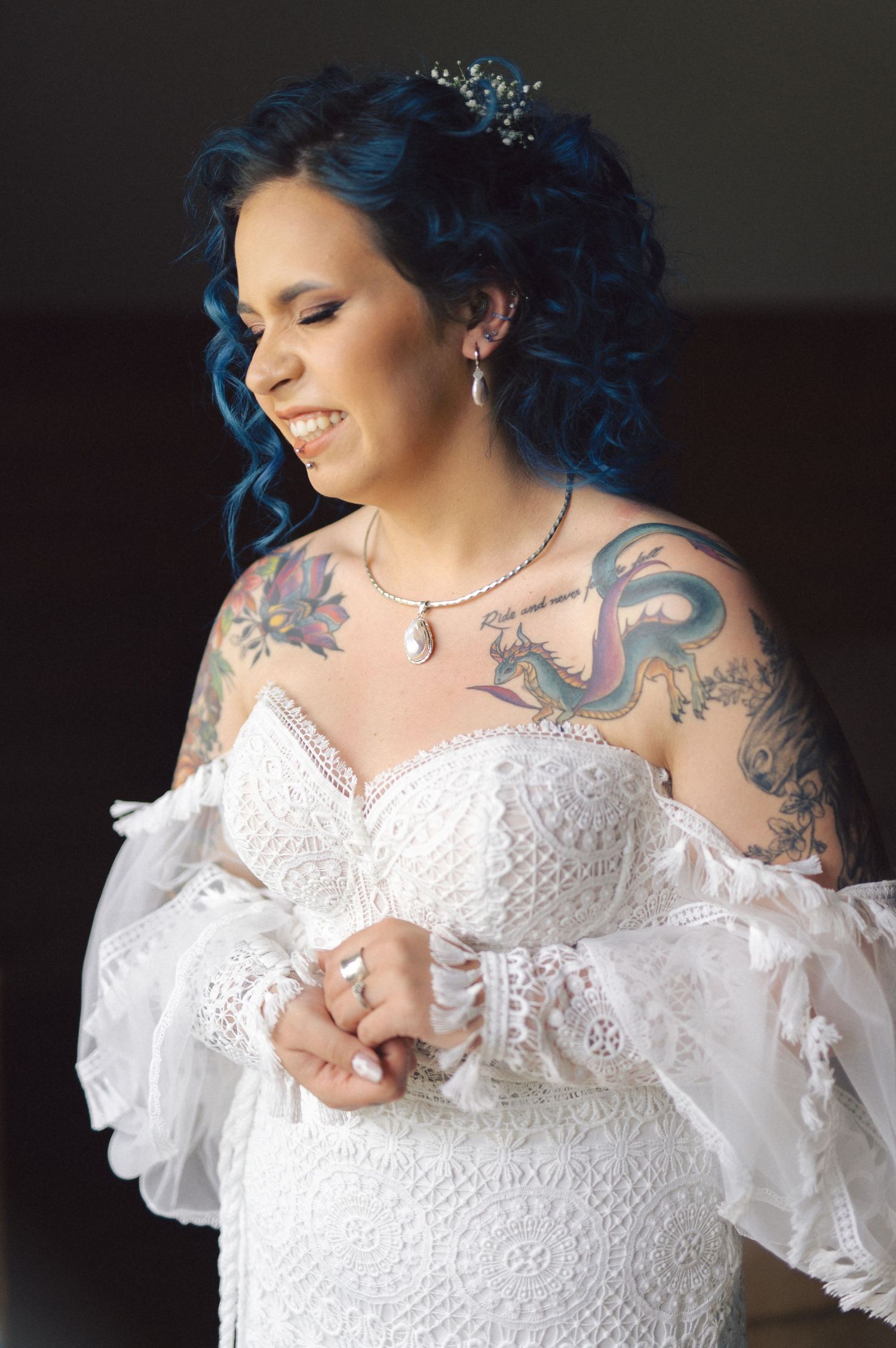 A bride in a white lace wedding dress with blue hair and visible tattoos smiles during a wedding photoshoot.