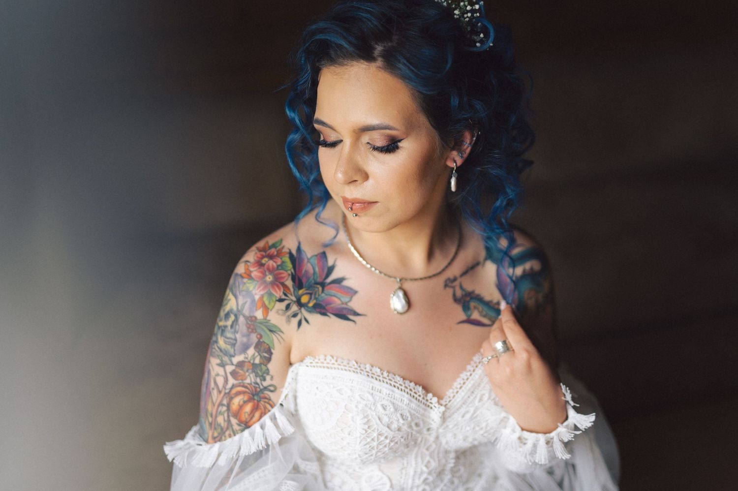 A bride in a lace wedding dress has blue hair and visible tattoos on her shoulder and arm.