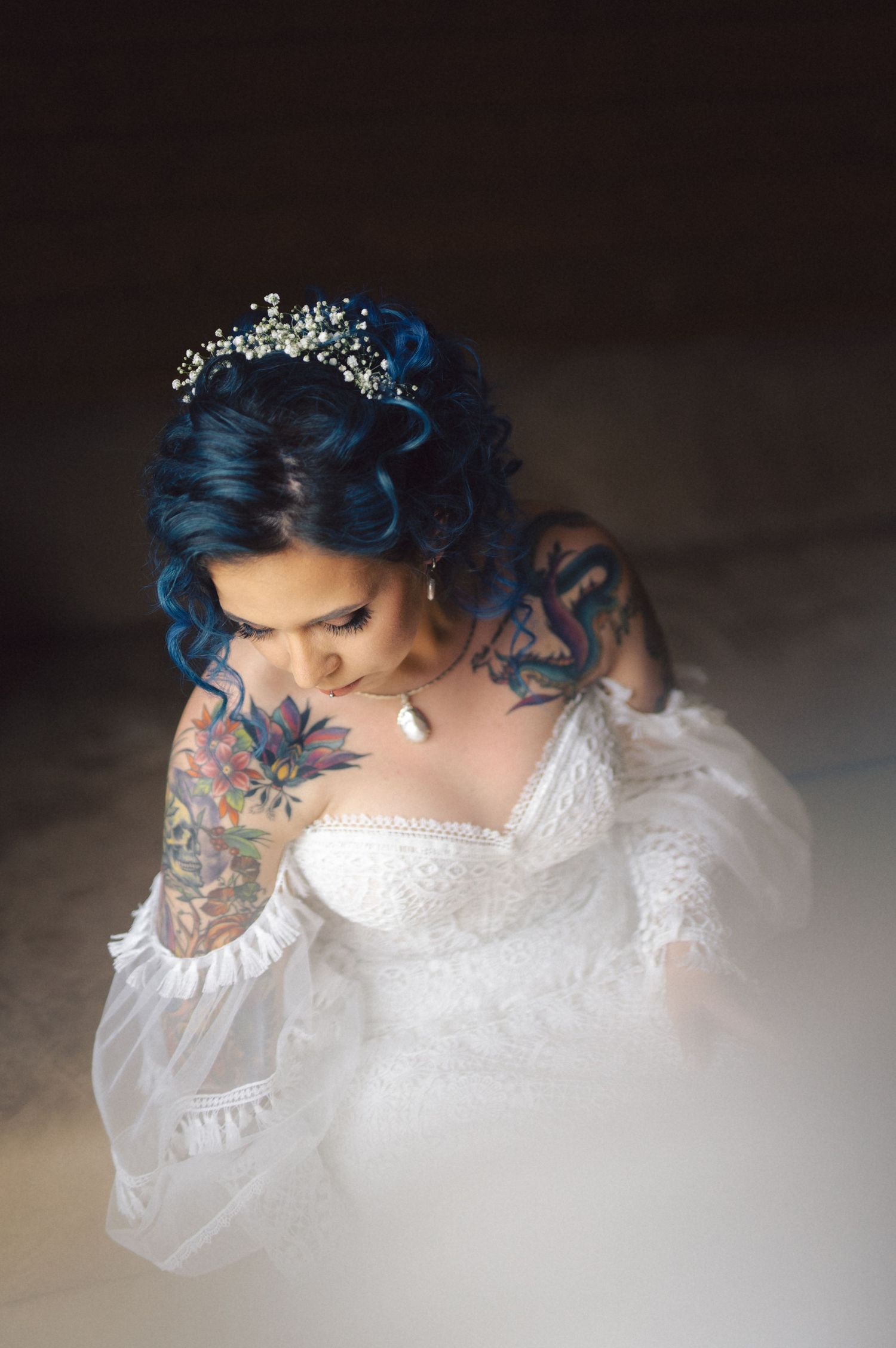 A bride in white lace dress with blue hair and arm tattoos looks down in a moody portrait shot.