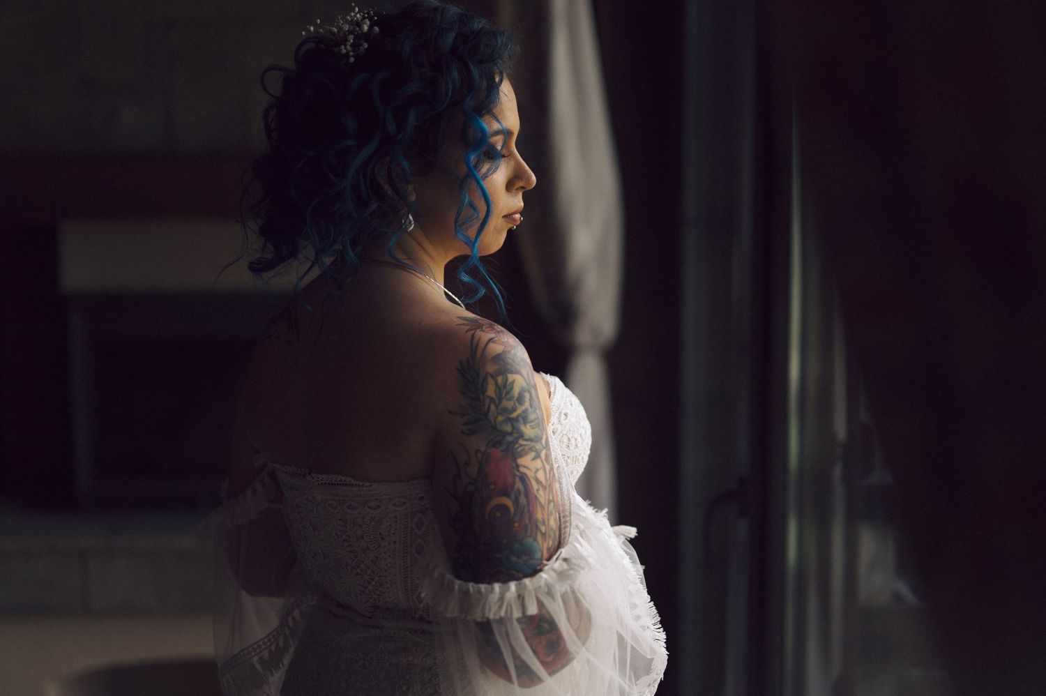 A bride in a white dress stands silhouetted against a window with dramatic lighting in a romantic wedding portrait.