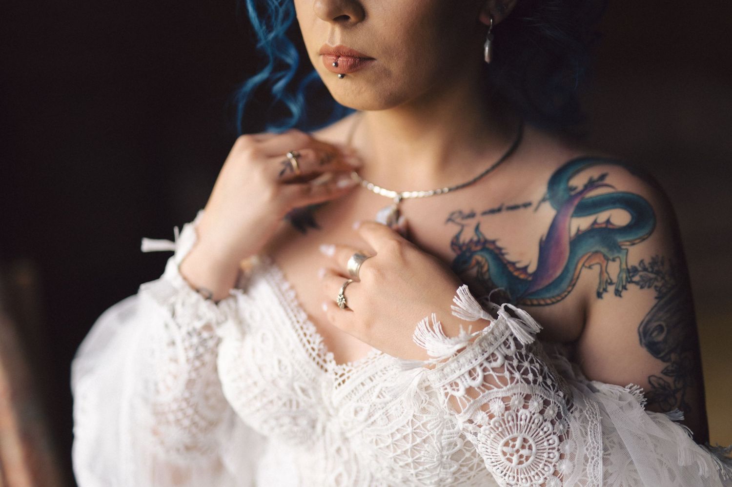 A person in white lace with visible blue dragon tattoos holds a smoking cigarette against a dark background.