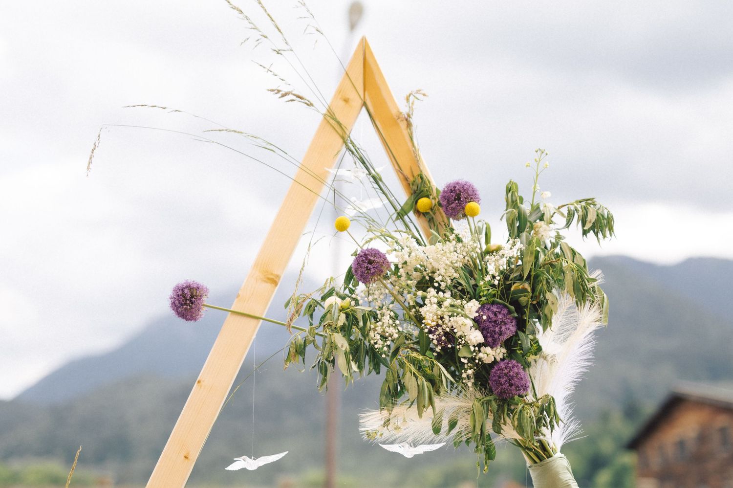 Triangle wooden wedding arch decorated with purple and white flowers against mountain backdrop in outdoor ceremony setting.