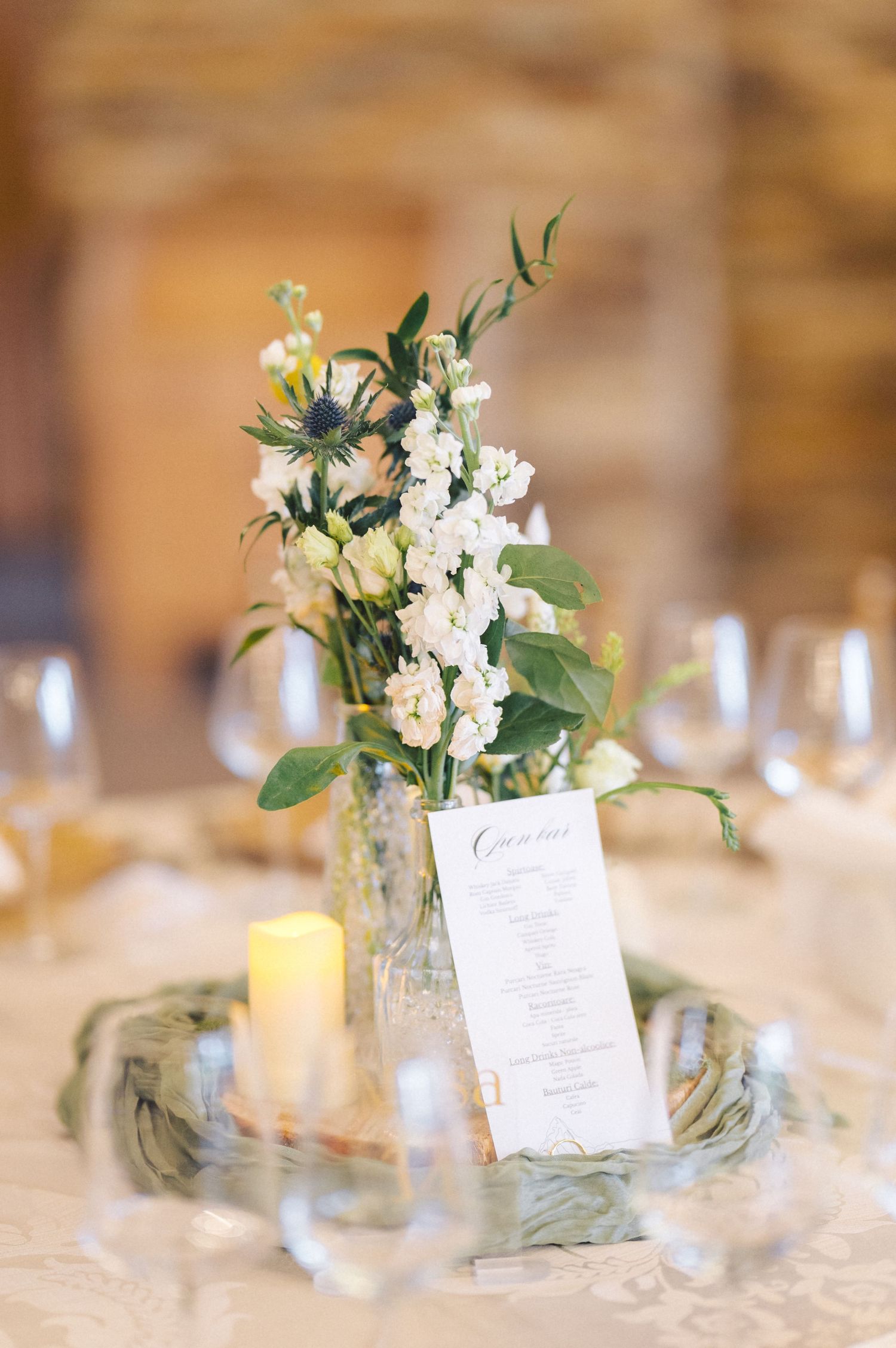 Elegant wedding table centerpiece with white flowers in a glass vase, menu card and glowing candle on lace tablecloth.