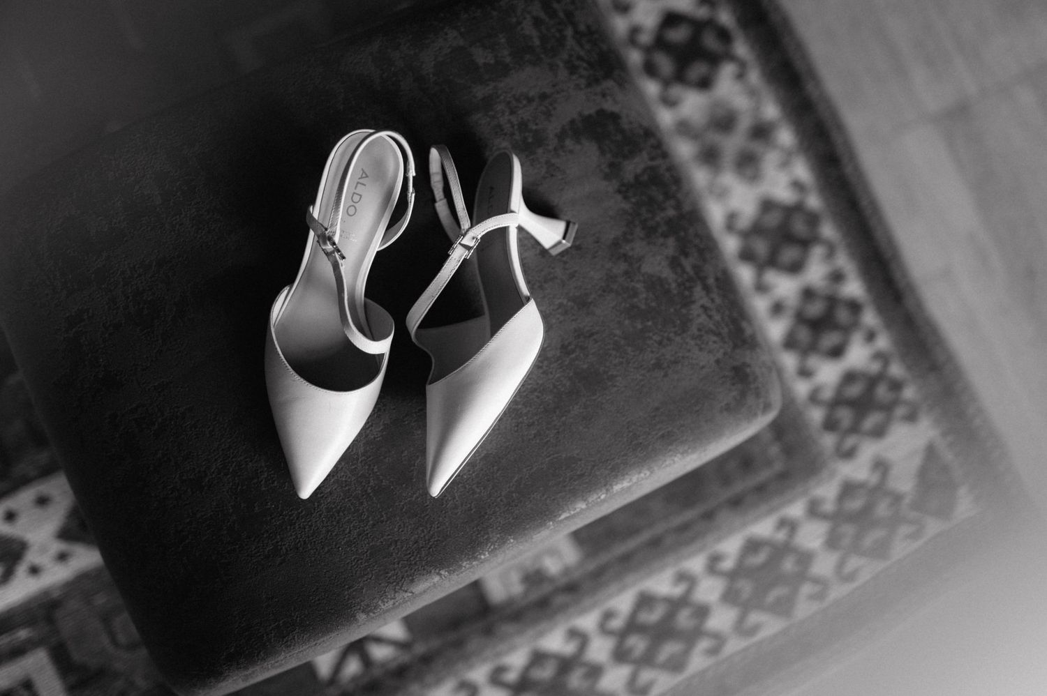 Elegant white pointed-toe slingback heels rest on a patterned rug in black and white photography.