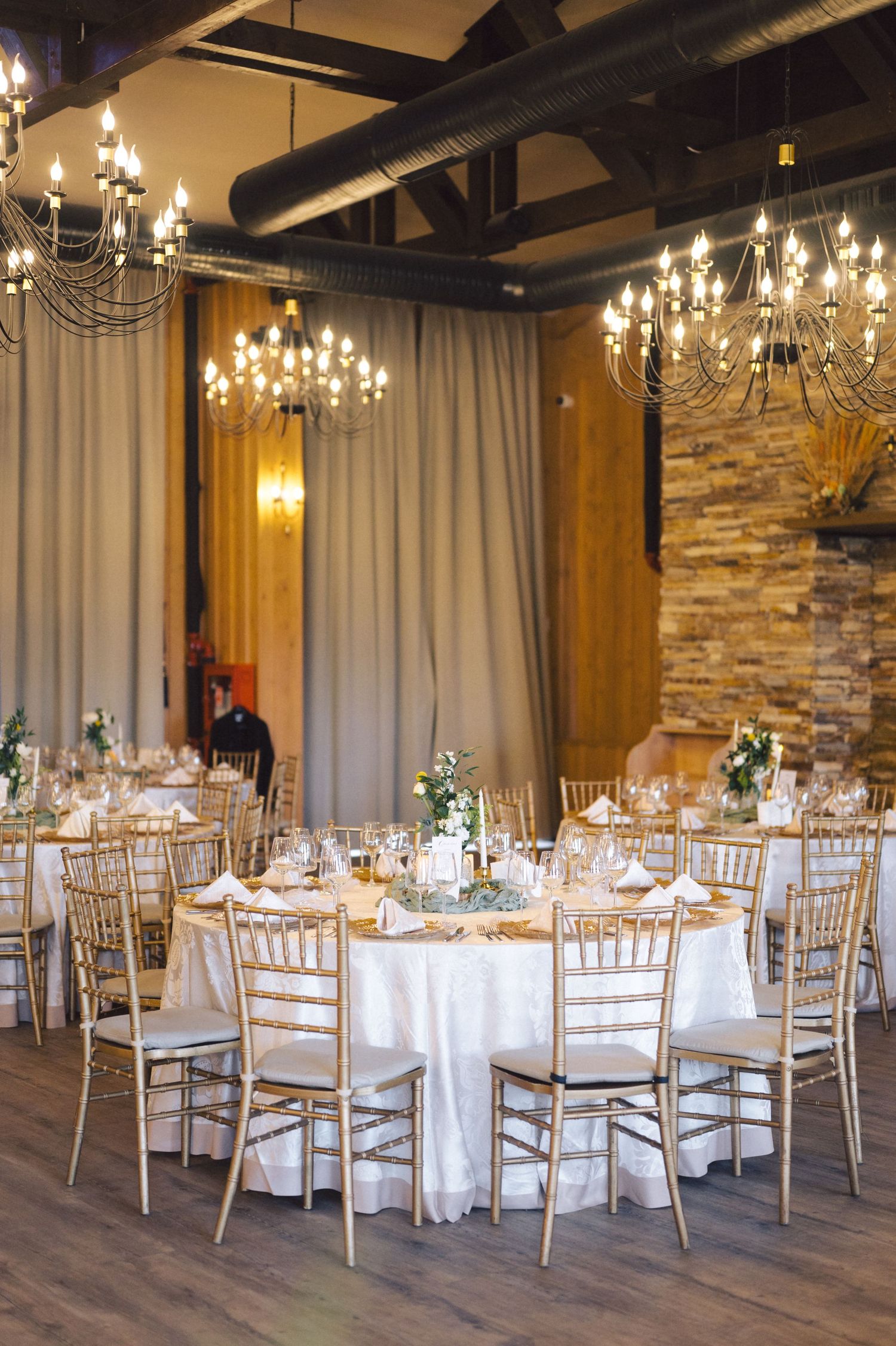 An elegant wedding reception venue with crystal chandeliers, gold chiavari chairs, and stone accent walls.