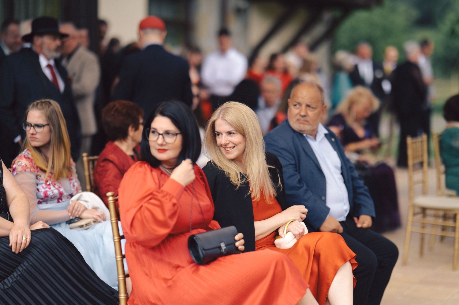 Guests seated at an outdoor event with people wearing formal attire including a vibrant orange dress.