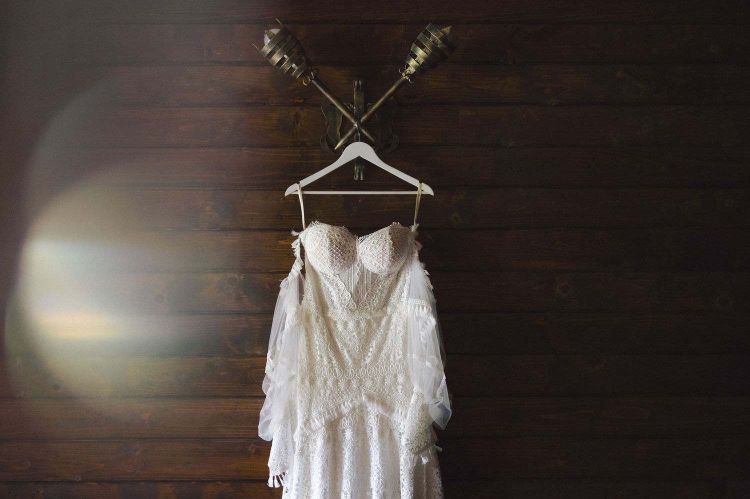 White lace wedding dress hanging on wooden hangers against dark wooden wall panels in soft lighting.