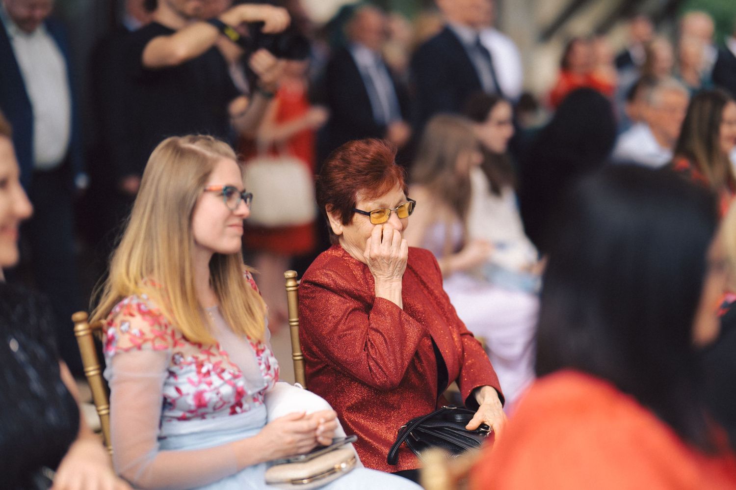 Two guests sit next to each other at an event, with one wearing a floral dress and another in a red outfit.