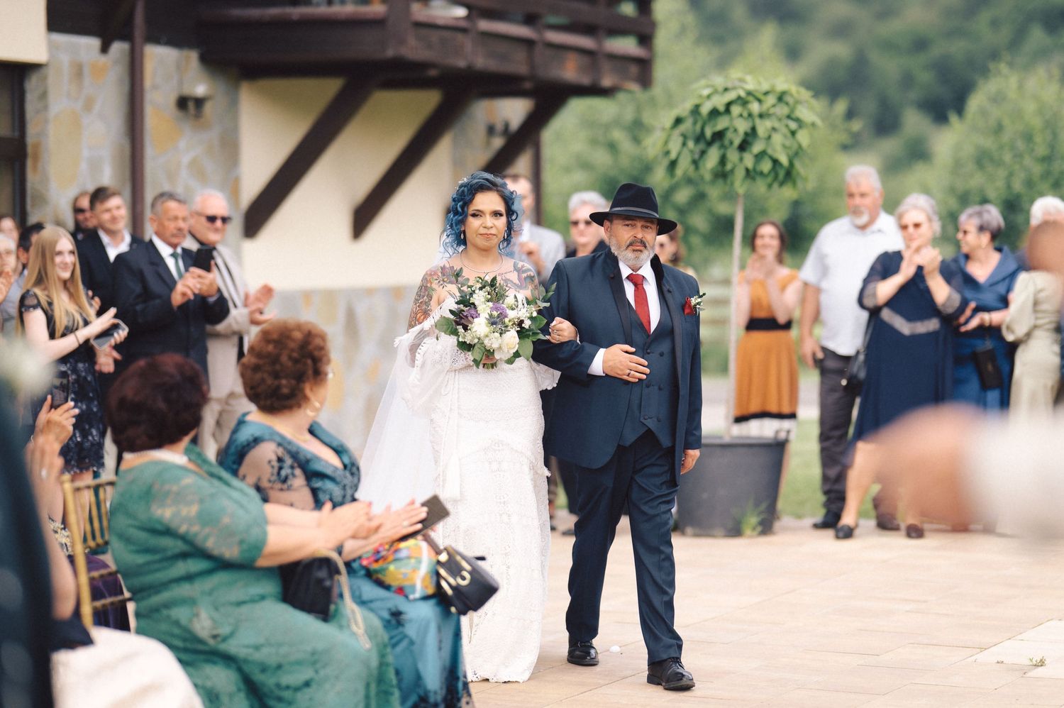 A couple walks down the aisle at an outdoor wedding ceremony while guests cheer and take photos.