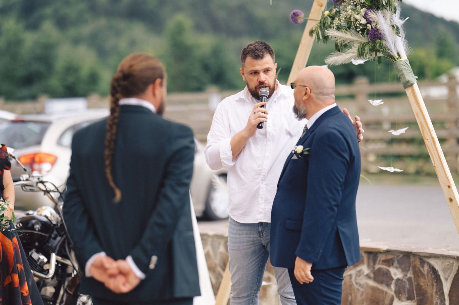 Wedding officiant speaking into microphone during outdoor lakeside ceremony under wooden arch.
