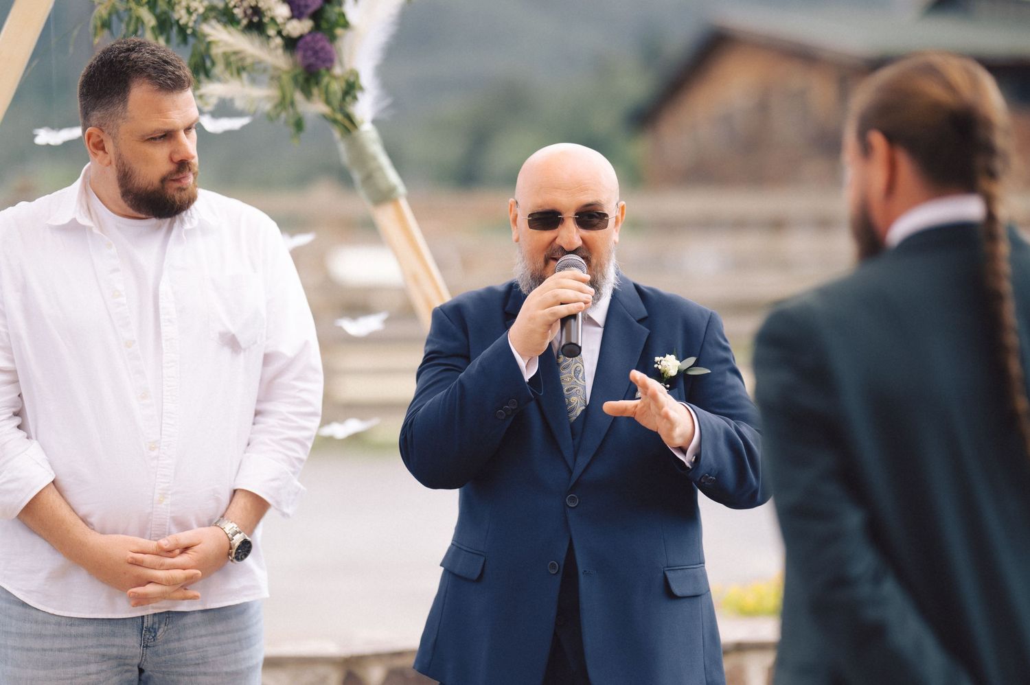 Three people interact during an outdoor wedding ceremony against a mountain backdrop.