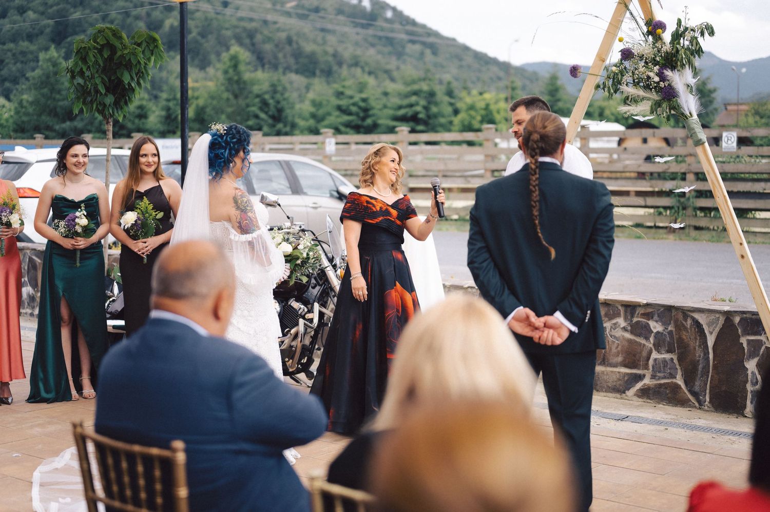 An outdoor wedding ceremony takes place under a wooden arch with mountain views and guests seated on rustic chairs.