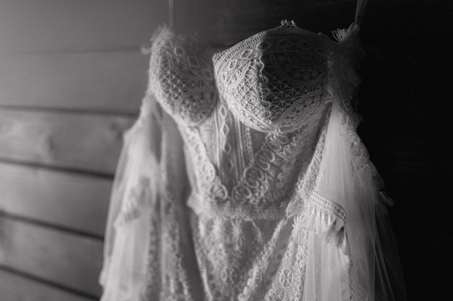 A delicate white lace wedding dress with sheer sleeves hangs against a wooden wall in black and white.