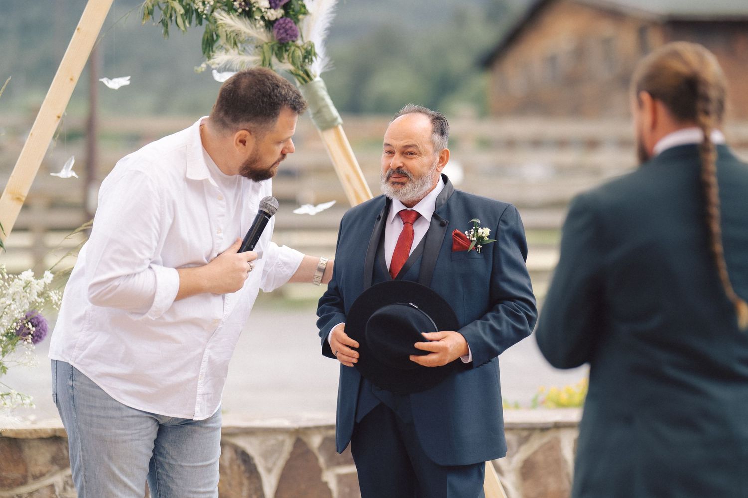 An outdoor wedding ceremony takes place under a floral arch with three people gathered on a stone terrace.