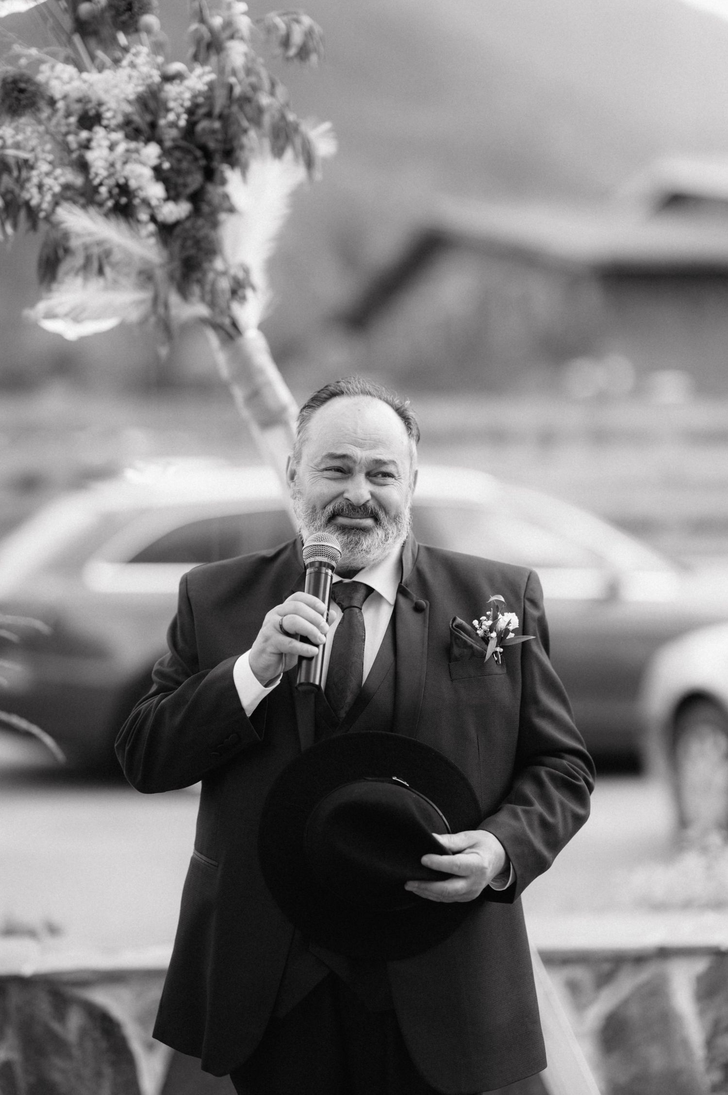 A person wearing a black suit speaks into a microphone while holding an item during an outdoor event.