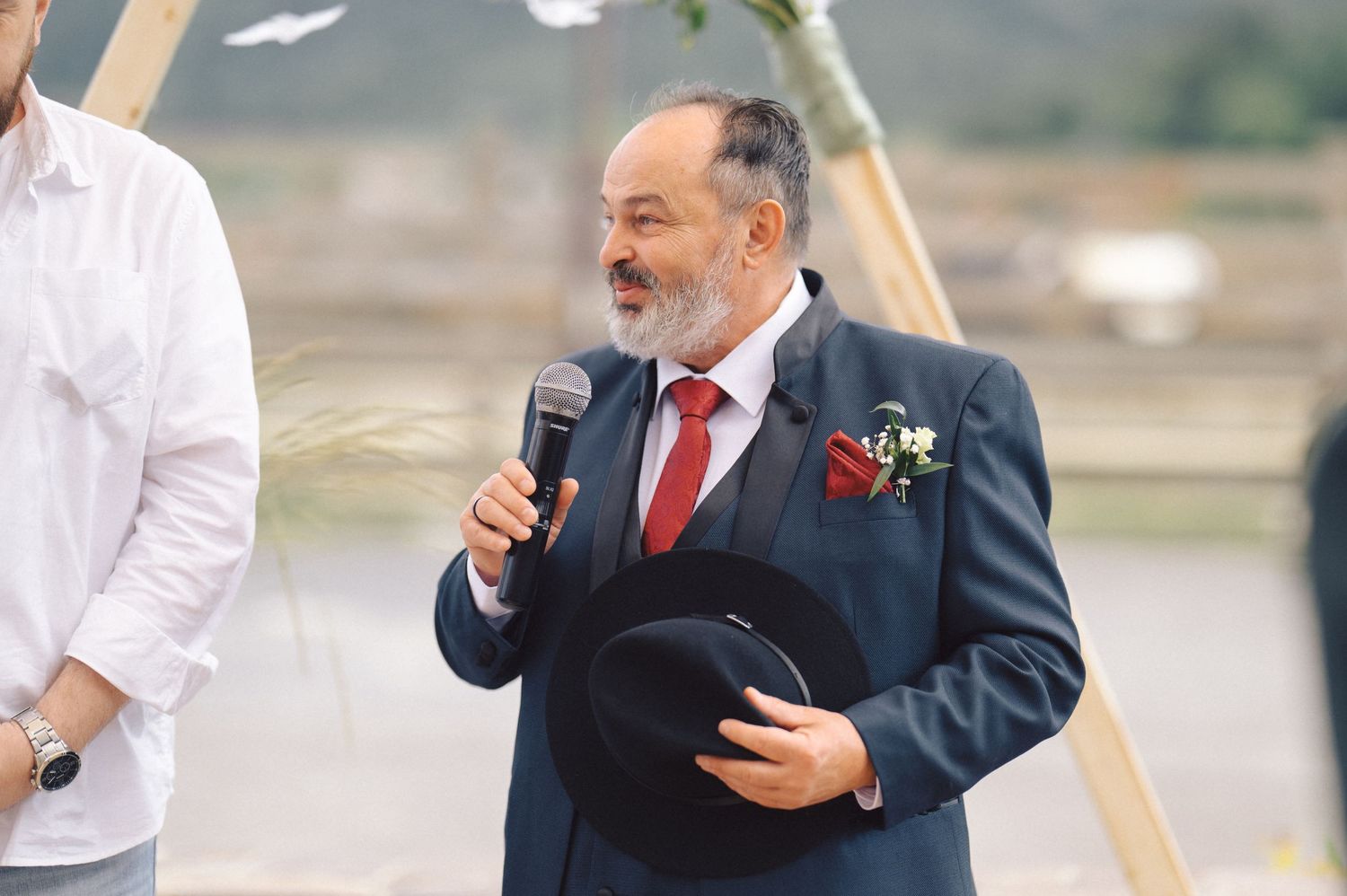 A speaker in a navy suit with red tie holds a microphone and book during an outdoor ceremony.