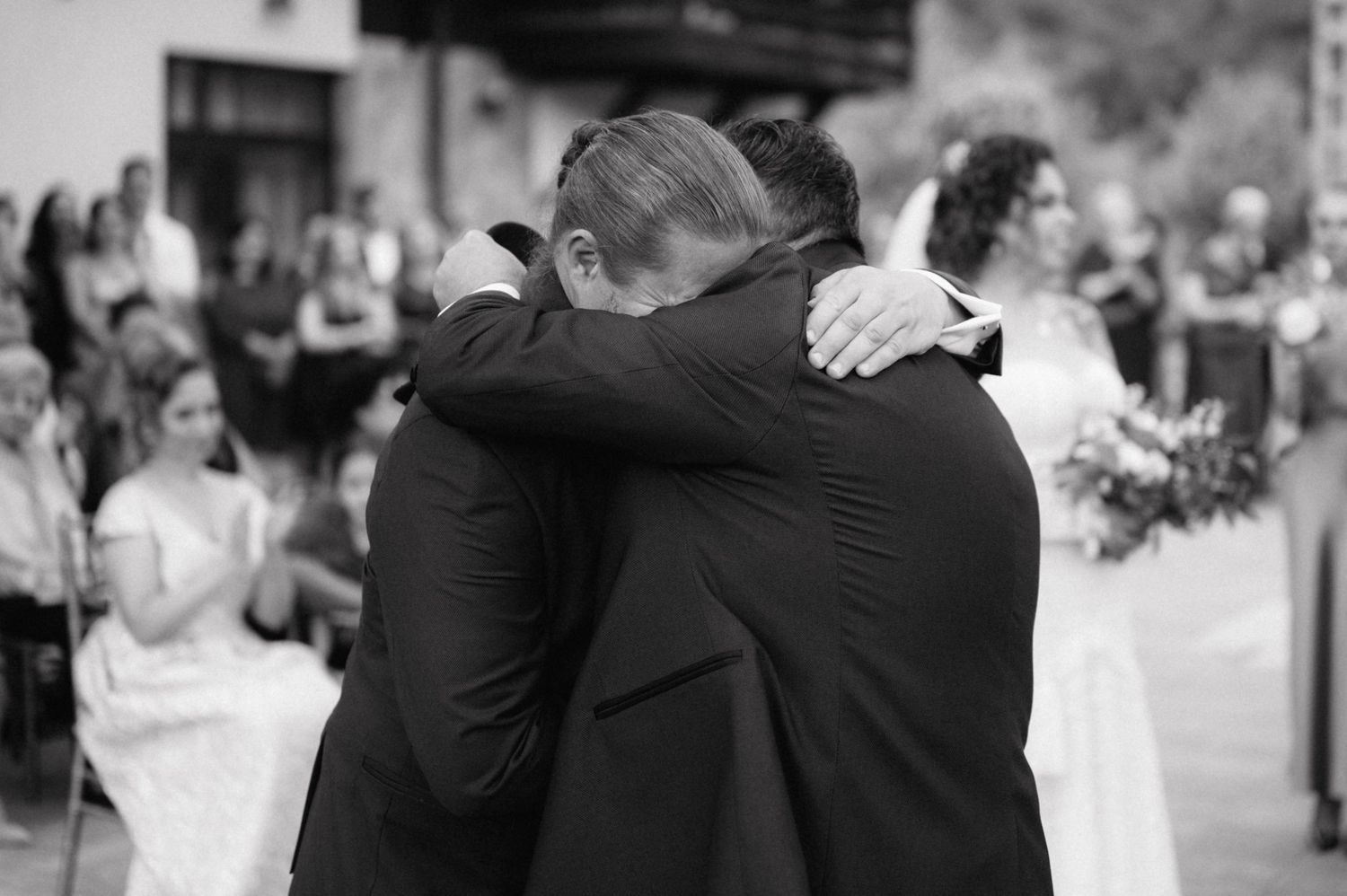 Two people embrace in a tender hug at a wedding celebration while guests look on in the background.