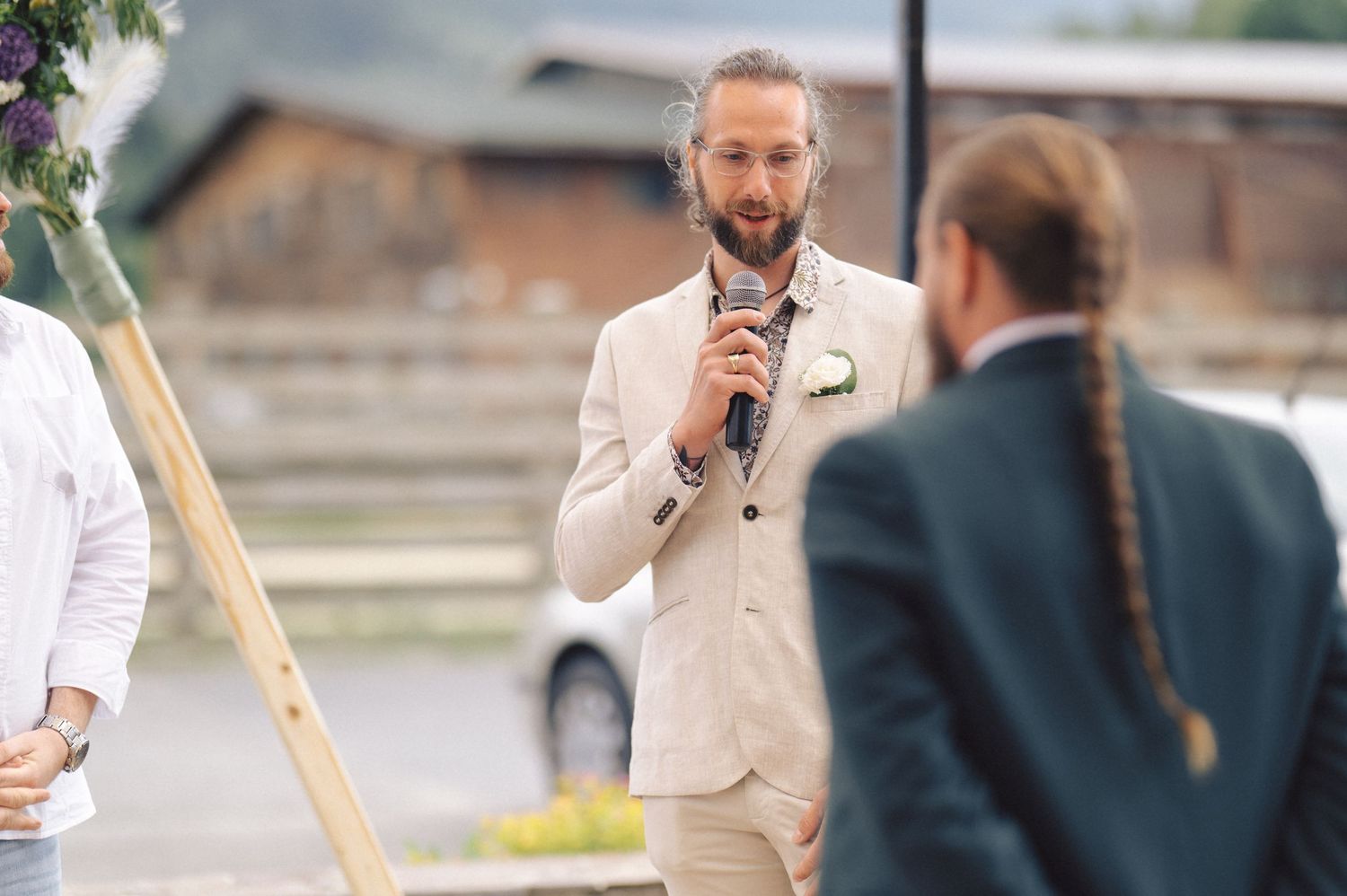 A speaker in a cream colored suit delivers a speech during an outdoor wedding ceremony.
