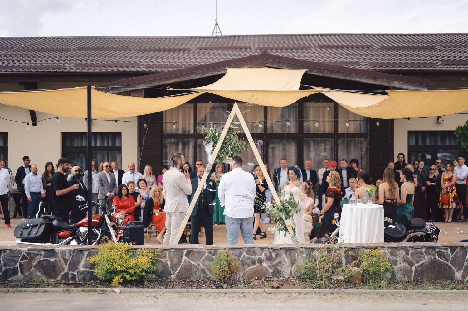 A garden wedding ceremony setup with triangular arch and guests gathered under cream-colored canopy shade sails.