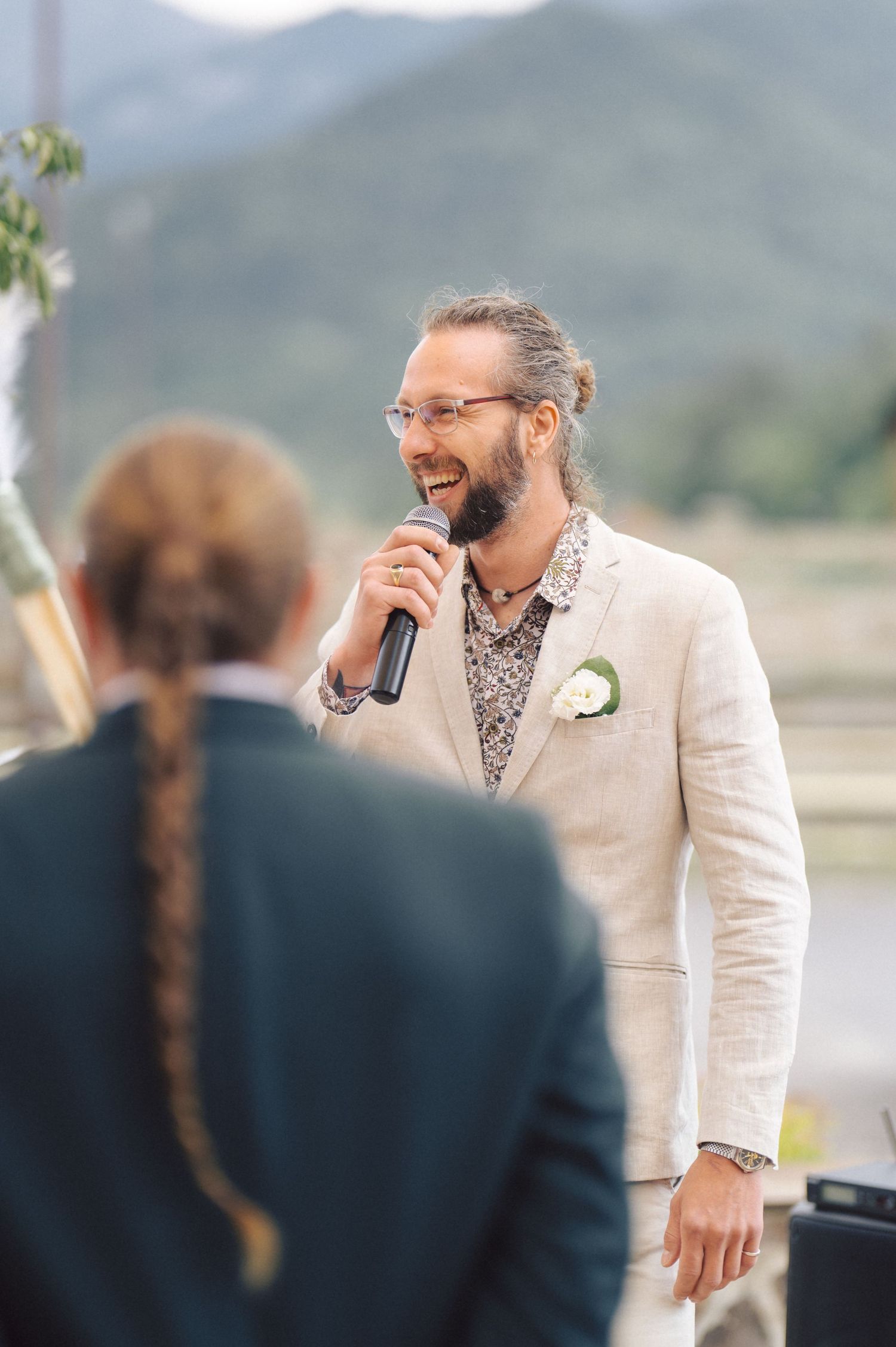 Groom wearing light linen suit and floral shirt speaks into microphone during outdoor wedding ceremony.