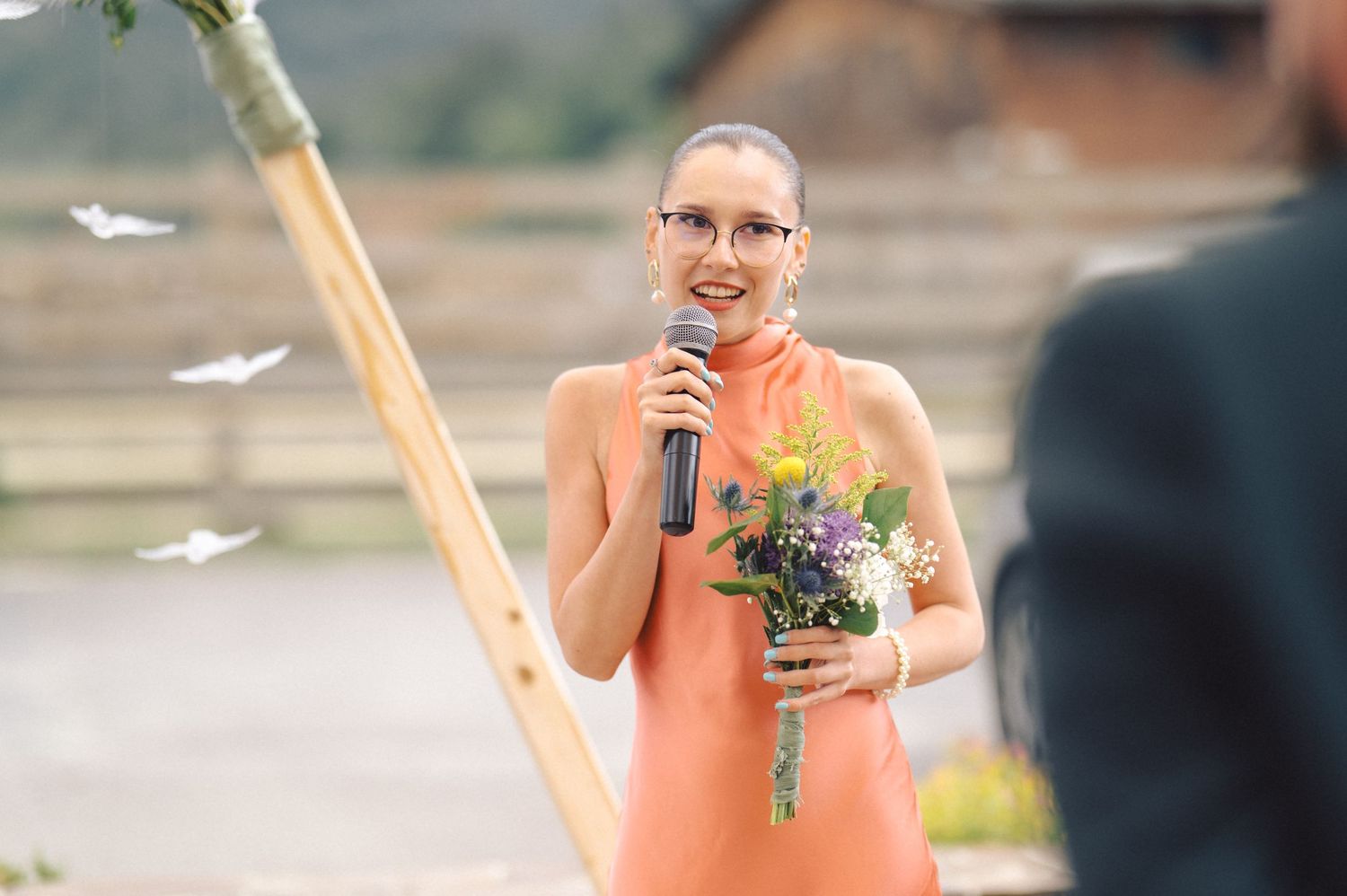 A person in a peach-colored dress holds a microphone and bouquet during an outdoor event while flower petals float in the air.