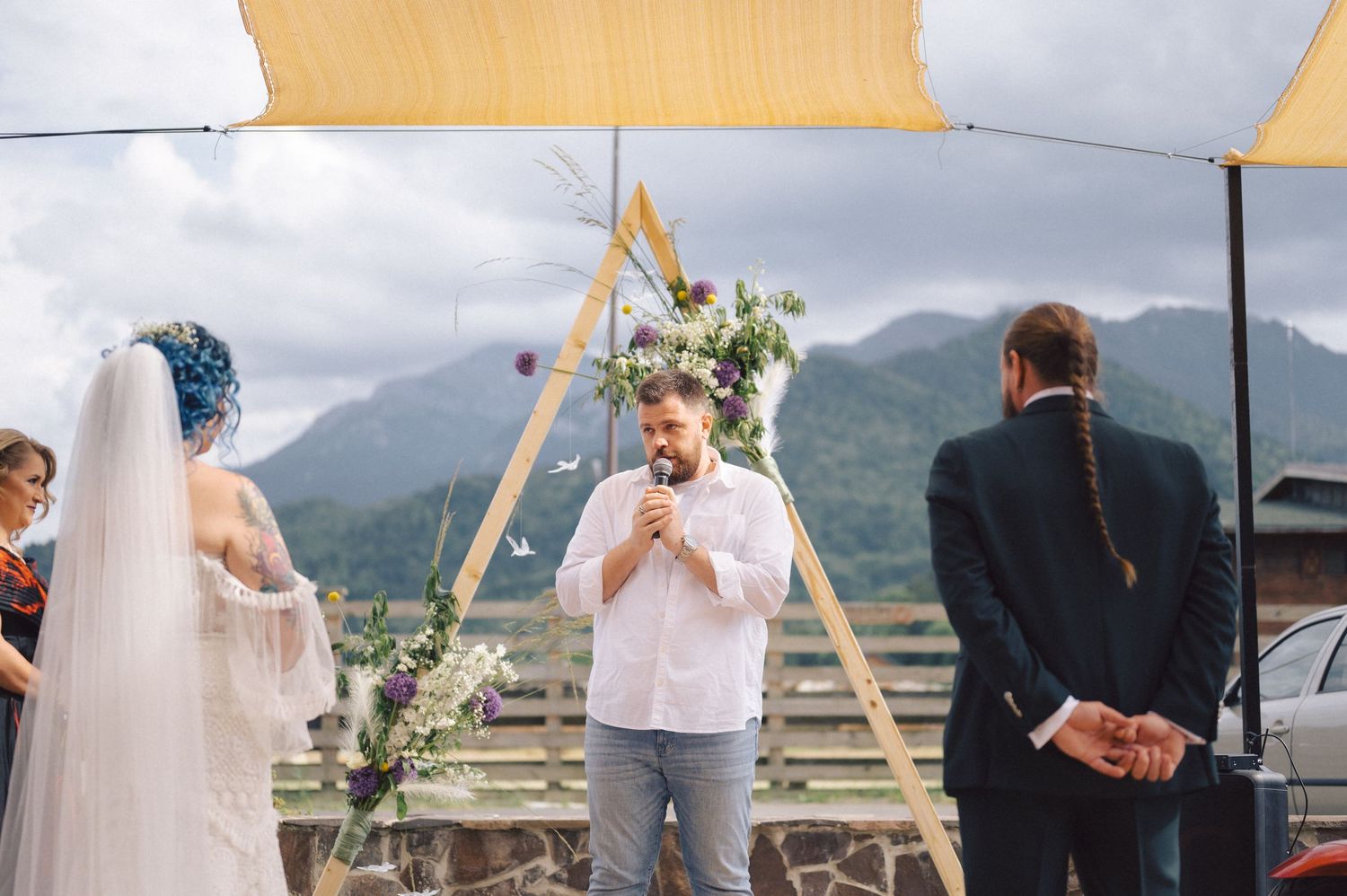 An outdoor wedding ceremony takes place against a backdrop of misty mountains, with a wooden triangular arch decorated with flowers.