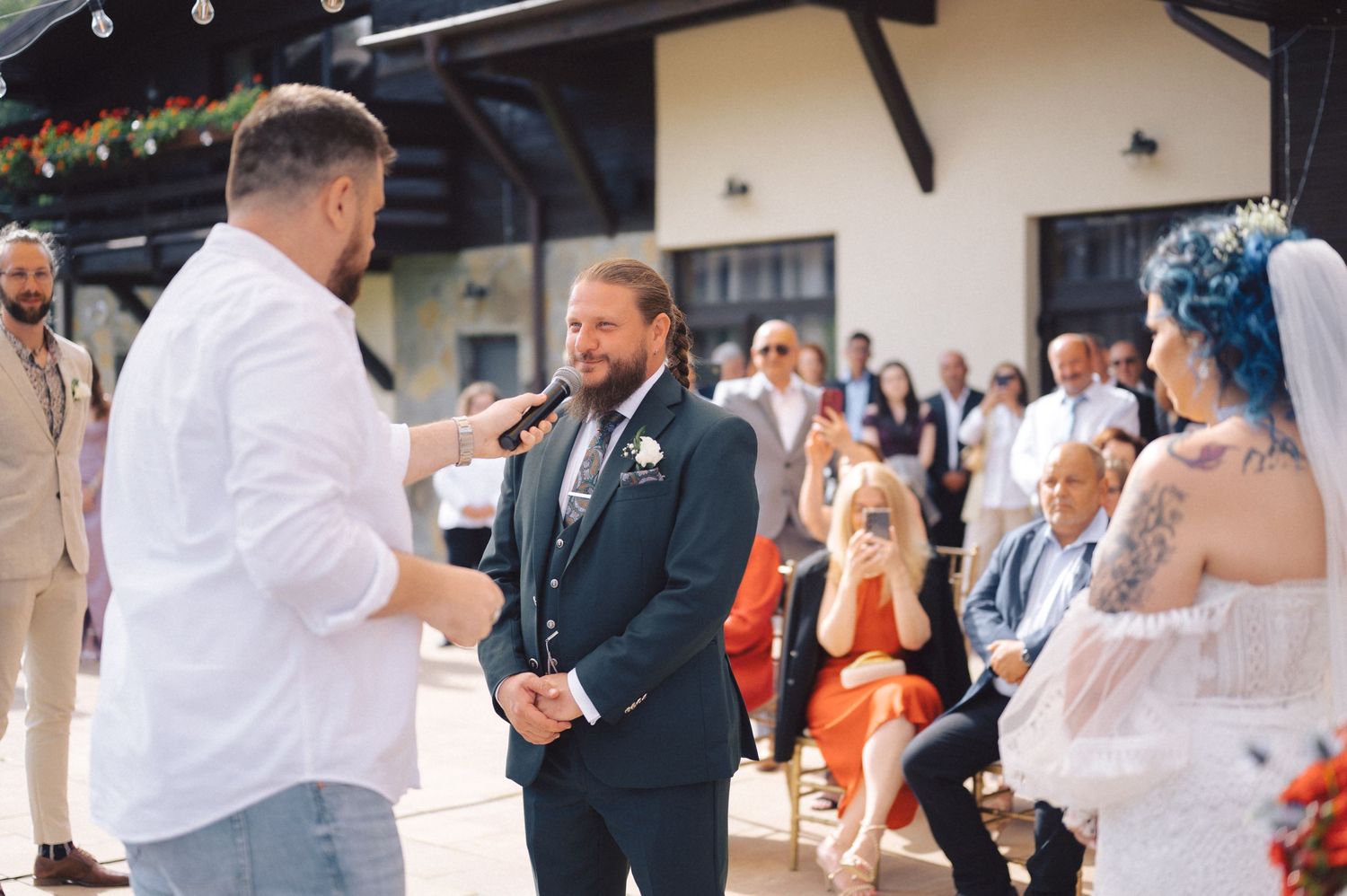 Wedding ceremony guests gather outdoors at a venue with string lights as someone in white speaks to someone in a suit.