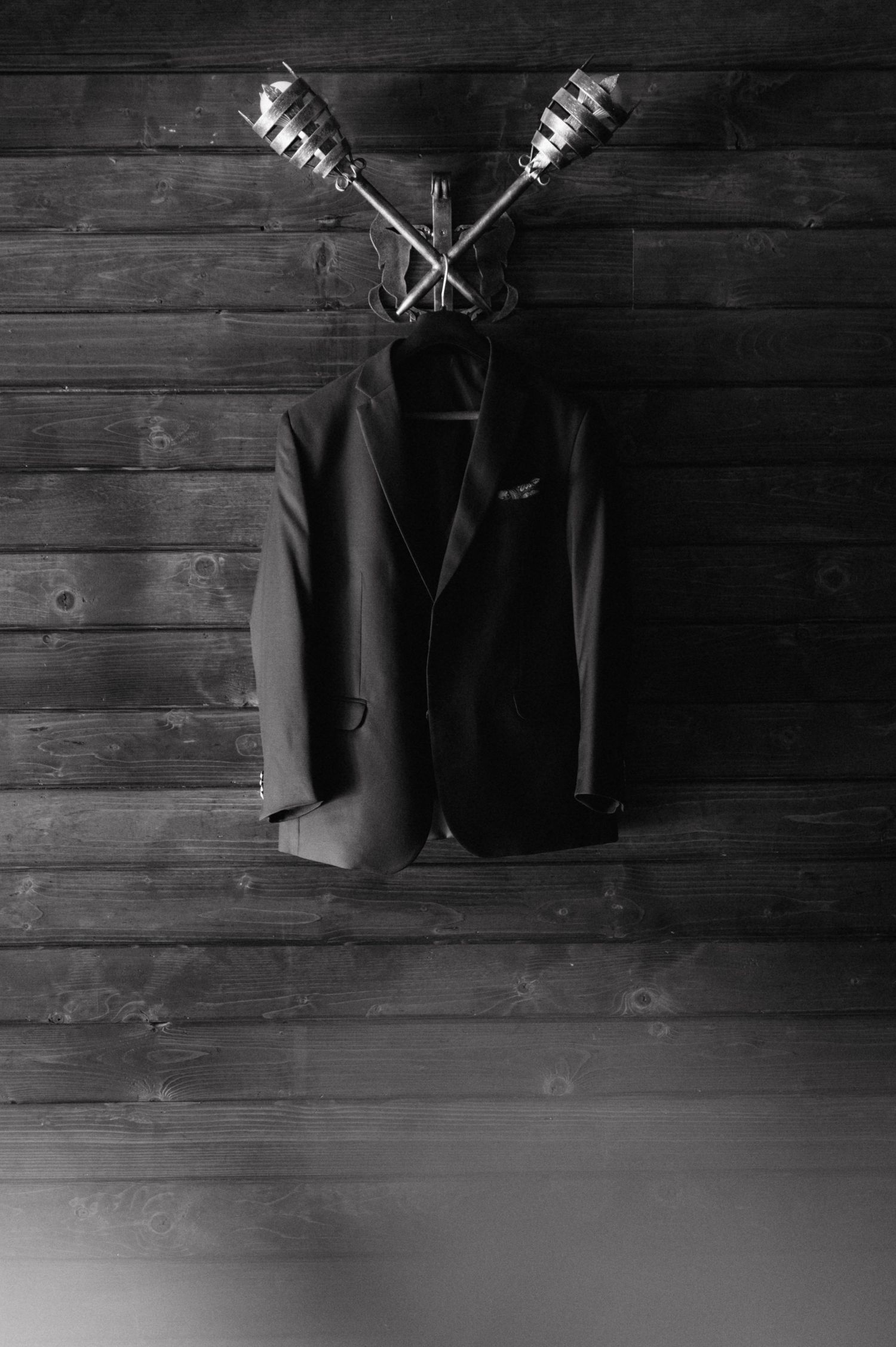 A black blazer jacket hangs on a wooden wall with two silver candlesticks mounted above it in a dark, moody setting.