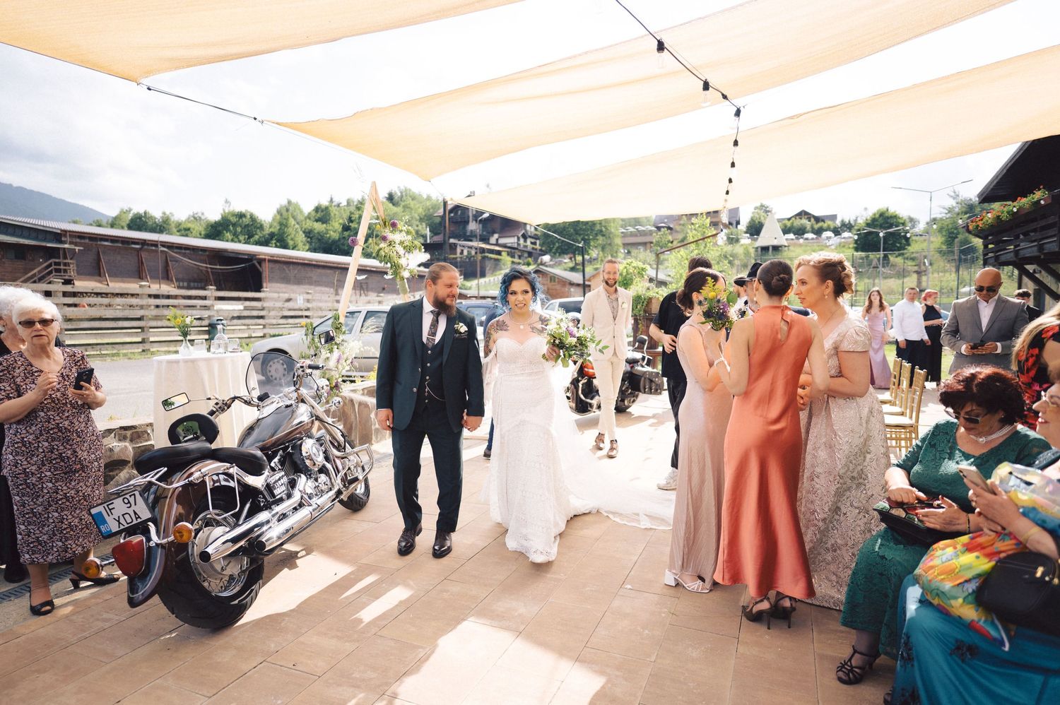 A happy couple walks together during a rustic outdoor wedding celebration near a parked motorcycle.