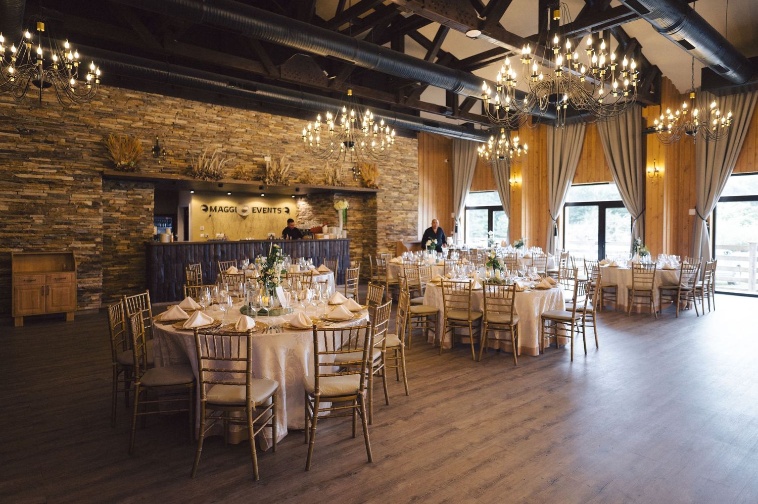 Rustic ballroom event space with stone walls, wooden beams, chandeliers, and round tables set for a formal dining occasion.