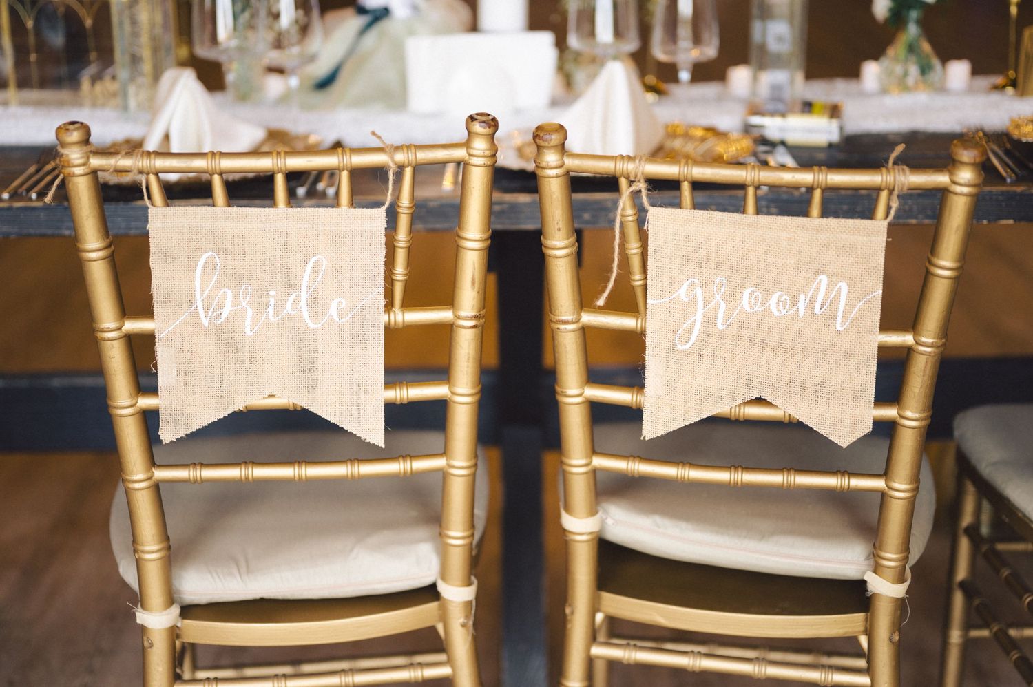 Gold wedding chairs decorated with vintage burlap banners reading 'bride' and 'groom' at an elegant reception setting.