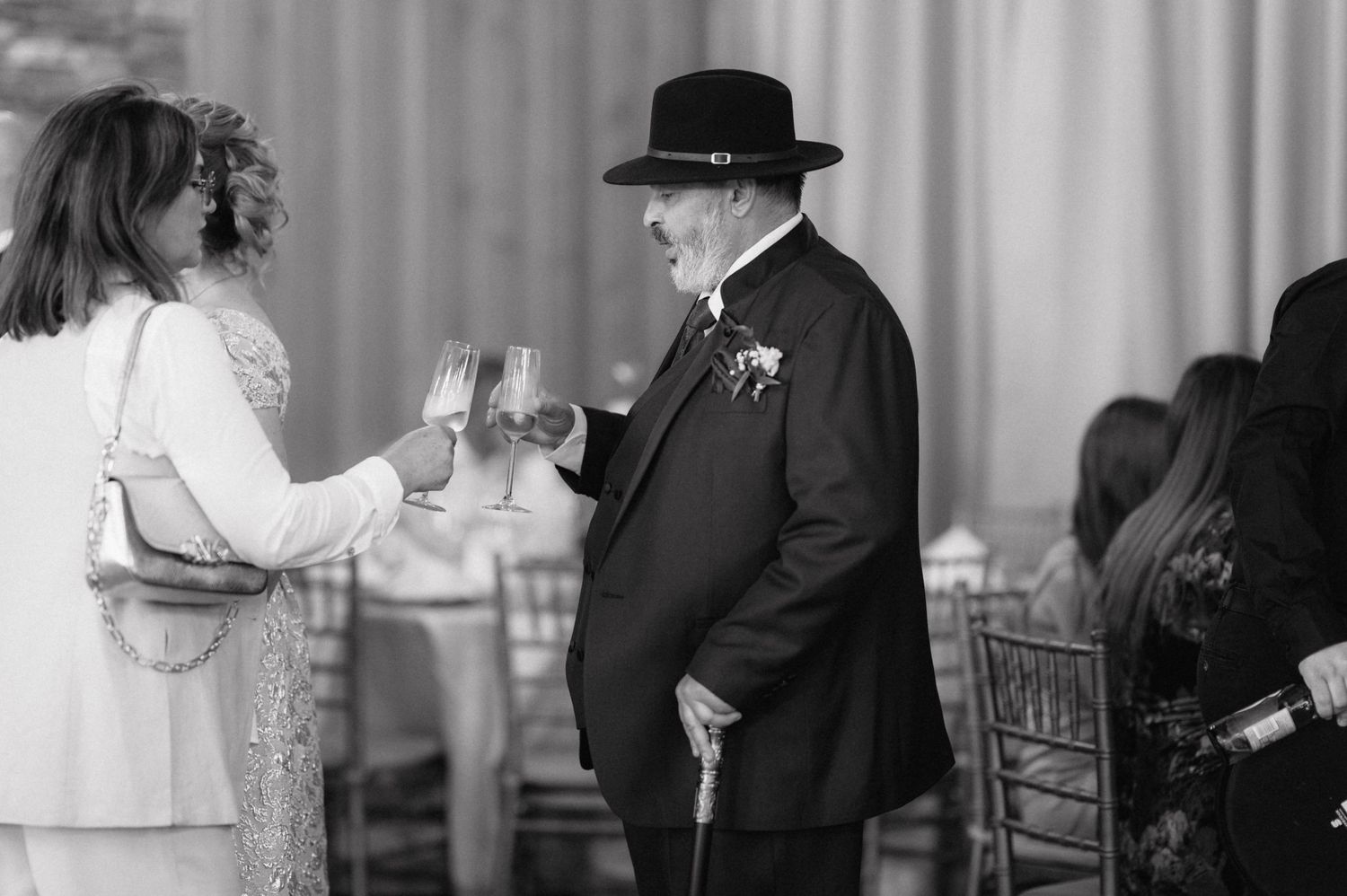 Black and white photo of two people sharing a toast at a formal event, with one wearing a dark suit and hat.