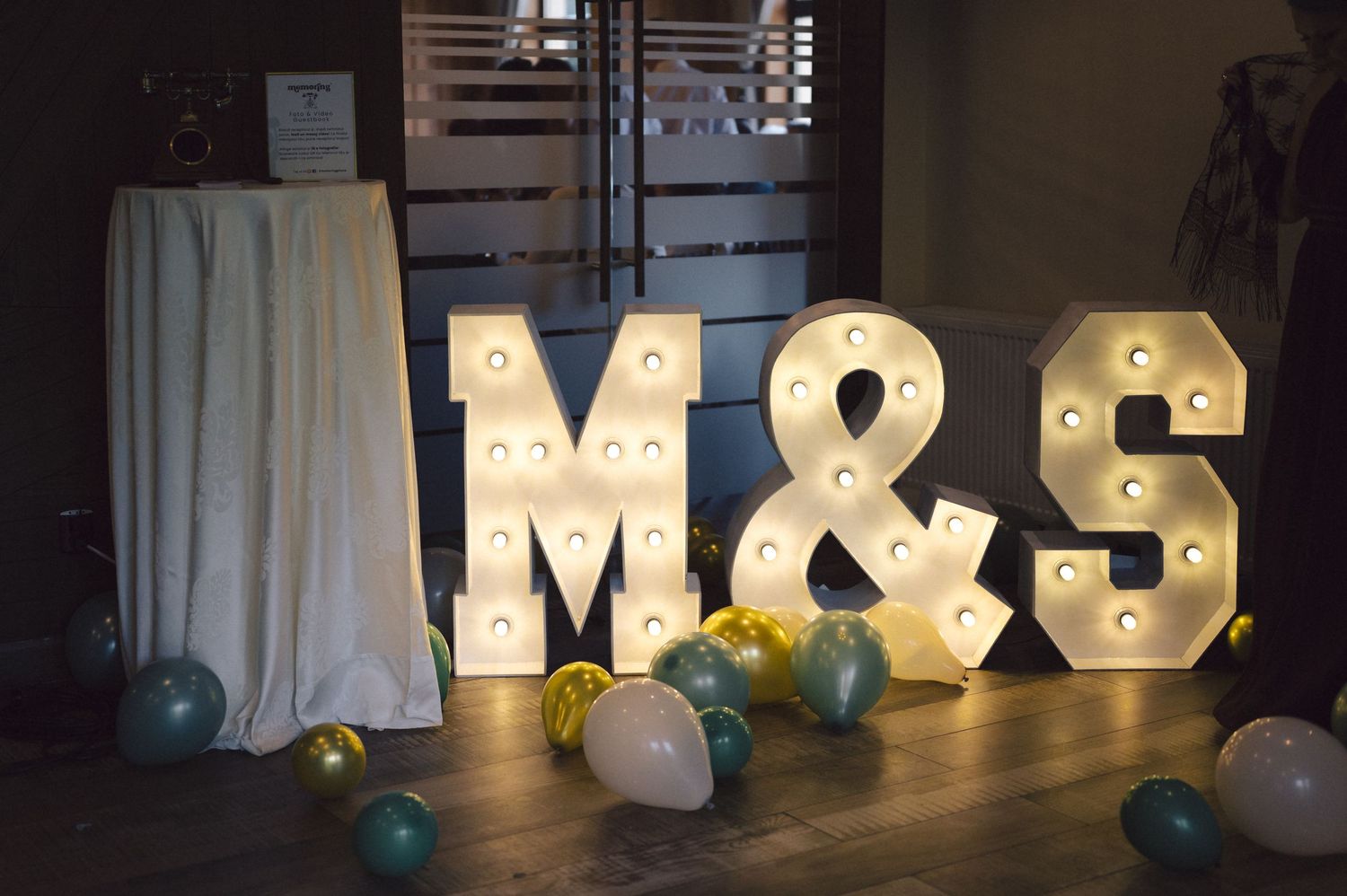 Large illuminated marquee letters M&S with ampersand stand glowing against dark background with scattered balloons.