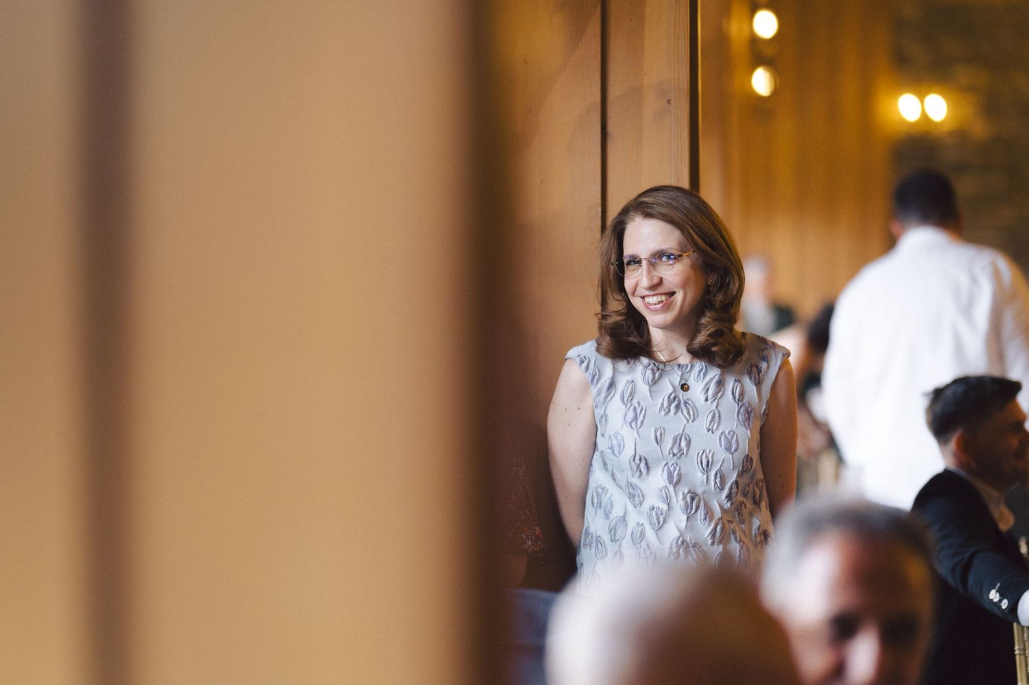 A person in a sleeveless lace dress stands among a crowd in a warmly lit indoor setting.