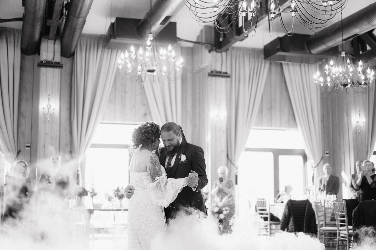 A couple shares their first dance in an elegant ballroom with chandeliers and white drapes during their wedding celebration.