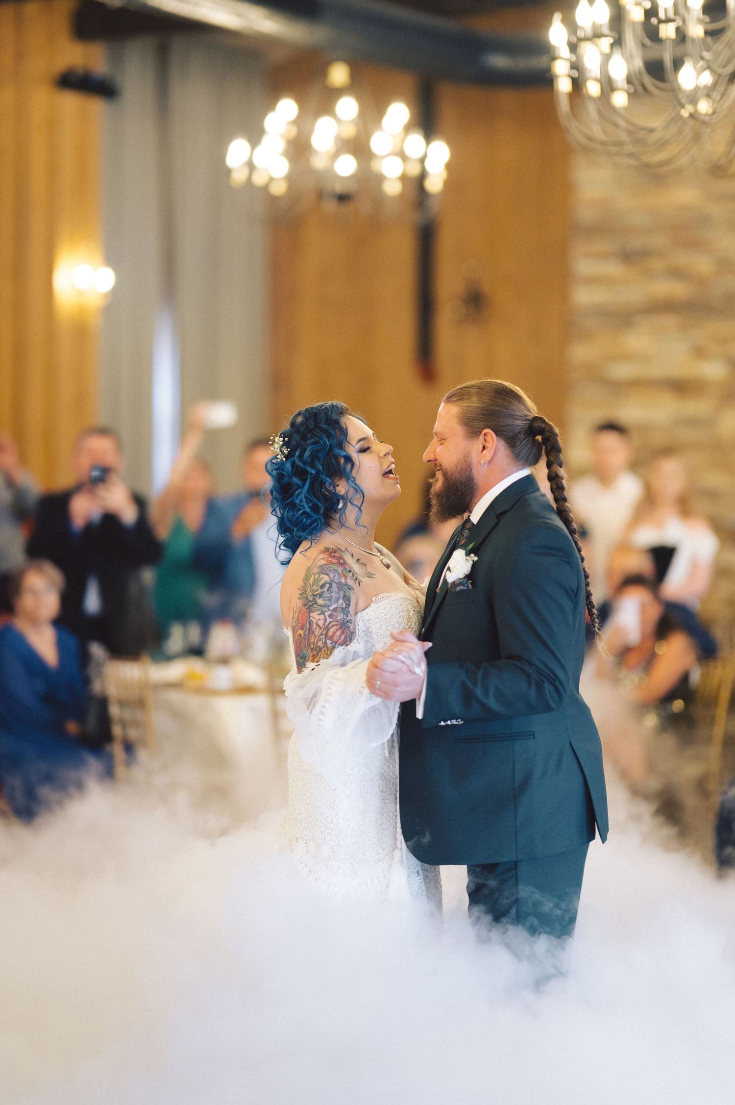 Newlywed couple shares their first dance on a misty dance floor under elegant chandeliers at indoor wedding reception.