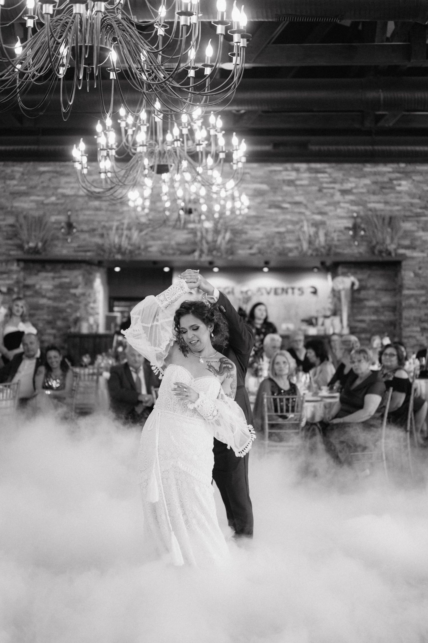 A couple shares their first dance on a misty dance floor beneath an elegant crystal chandelier in a rustic stone venue.