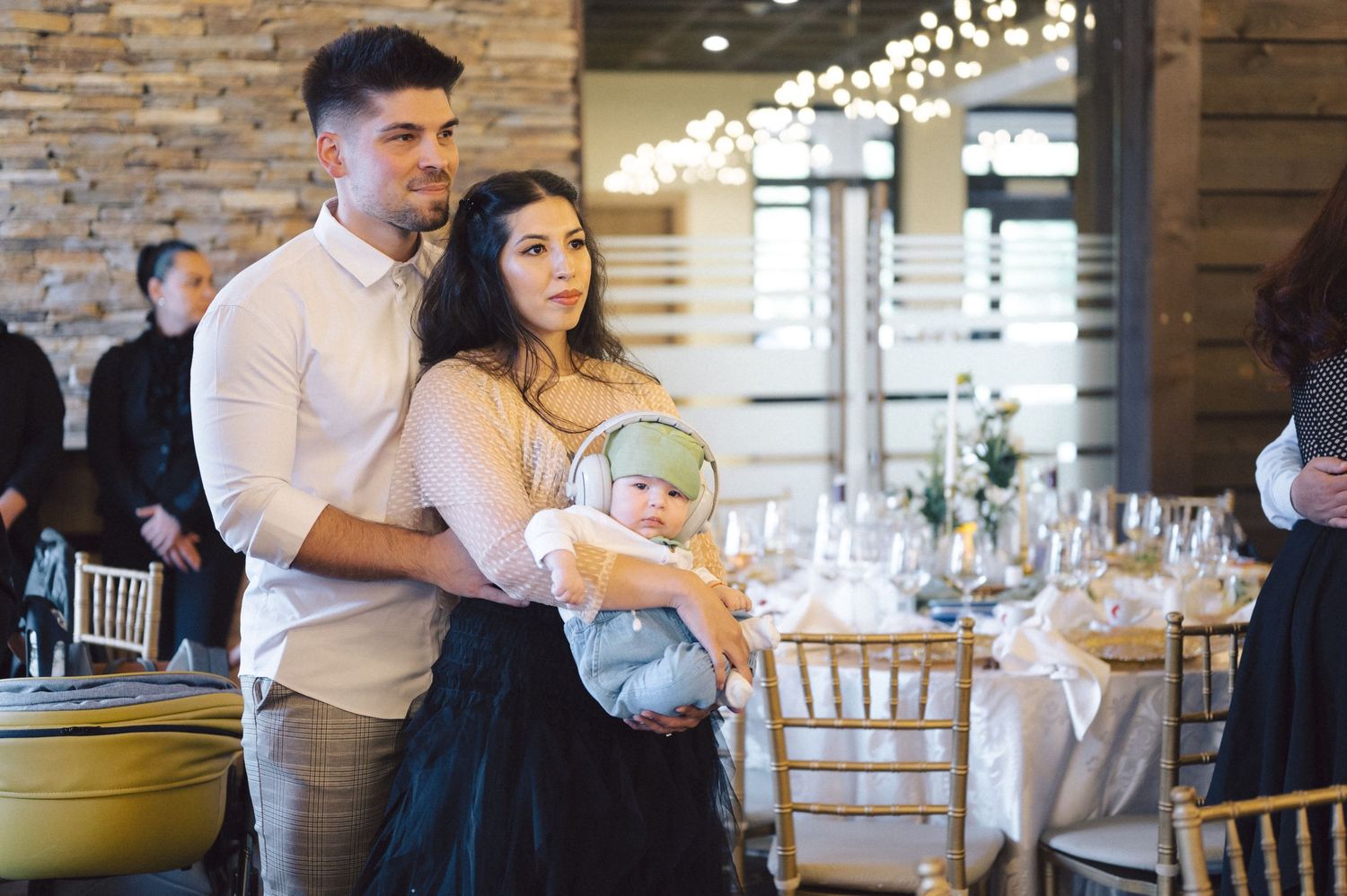 A couple holds their baby at an elegant reception venue with stone walls and table settings in the background.