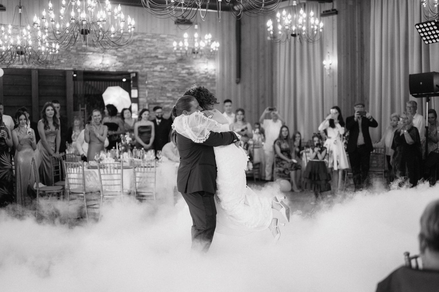 Newlyweds share their first dance on a fog-covered dance floor under crystal chandeliers at an elegant wedding reception.