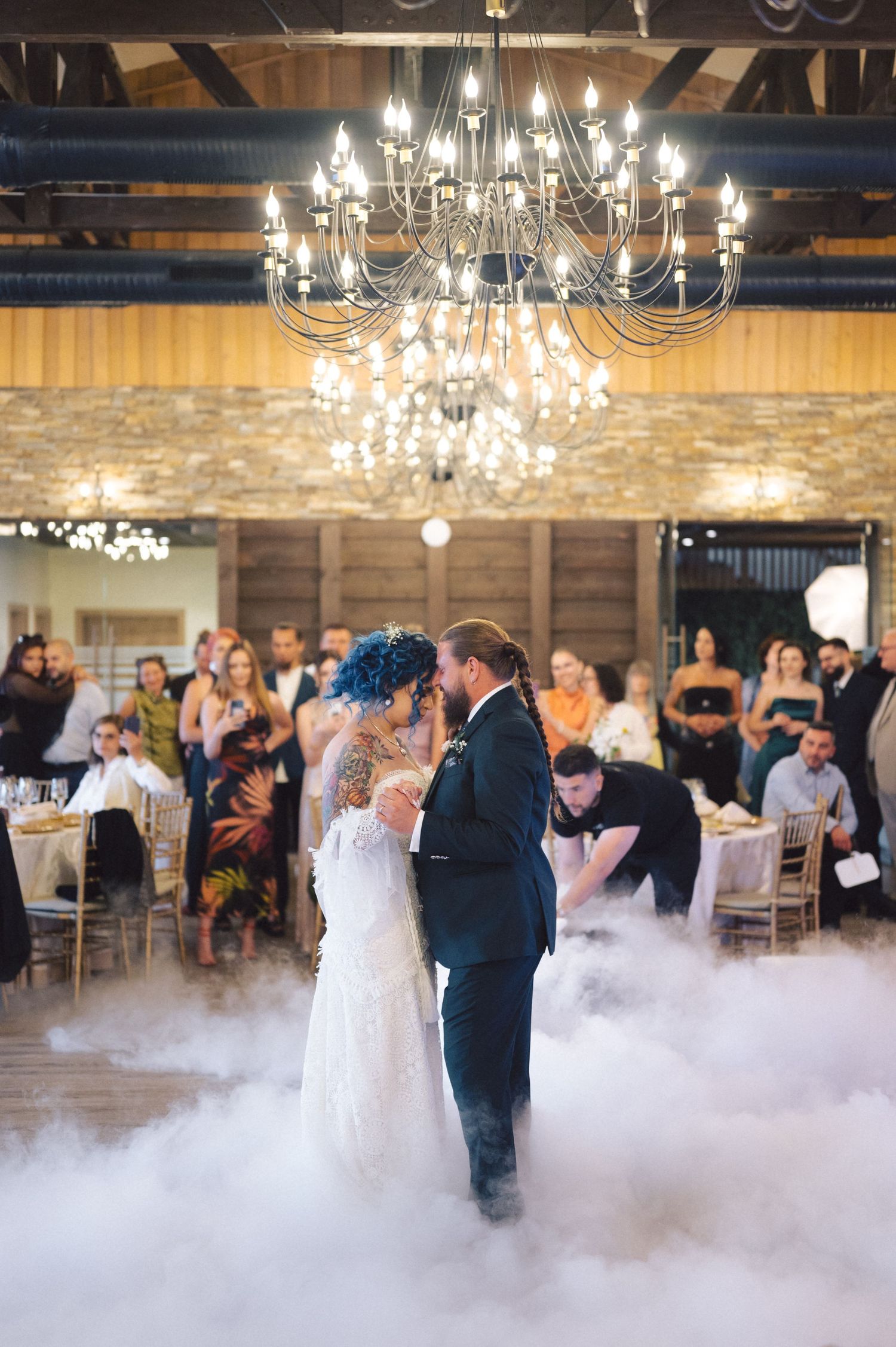 A couple shares their first dance at a rustic barn reception under a crystal chandelier with fog effects on the dance floor.
