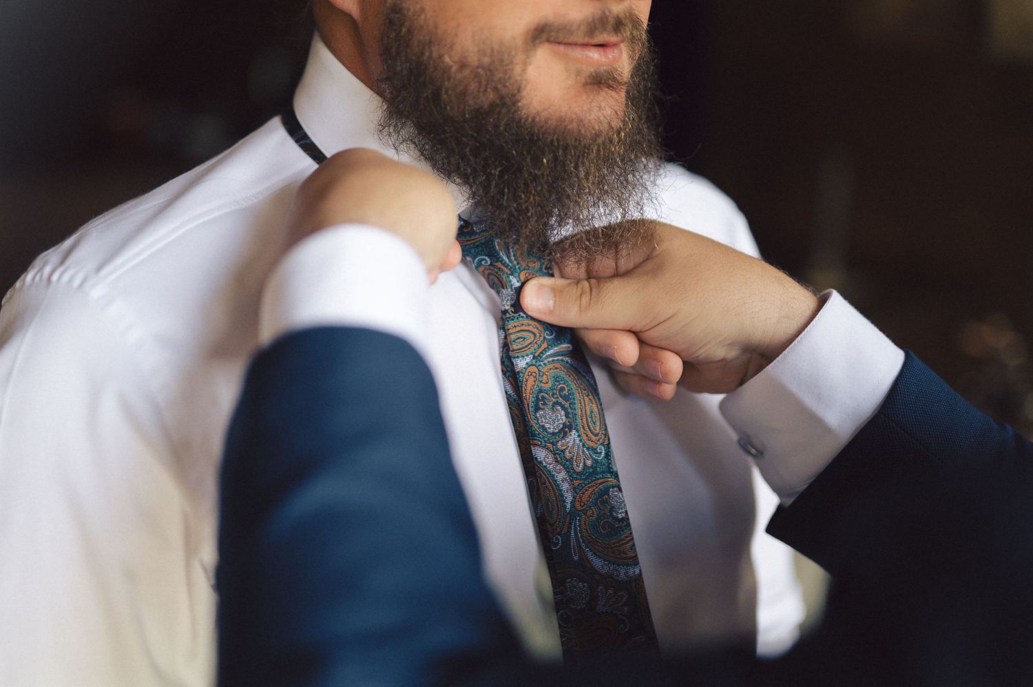 Close-up shot showing hands adjusting a patterned tie against a white shirt and navy suit.