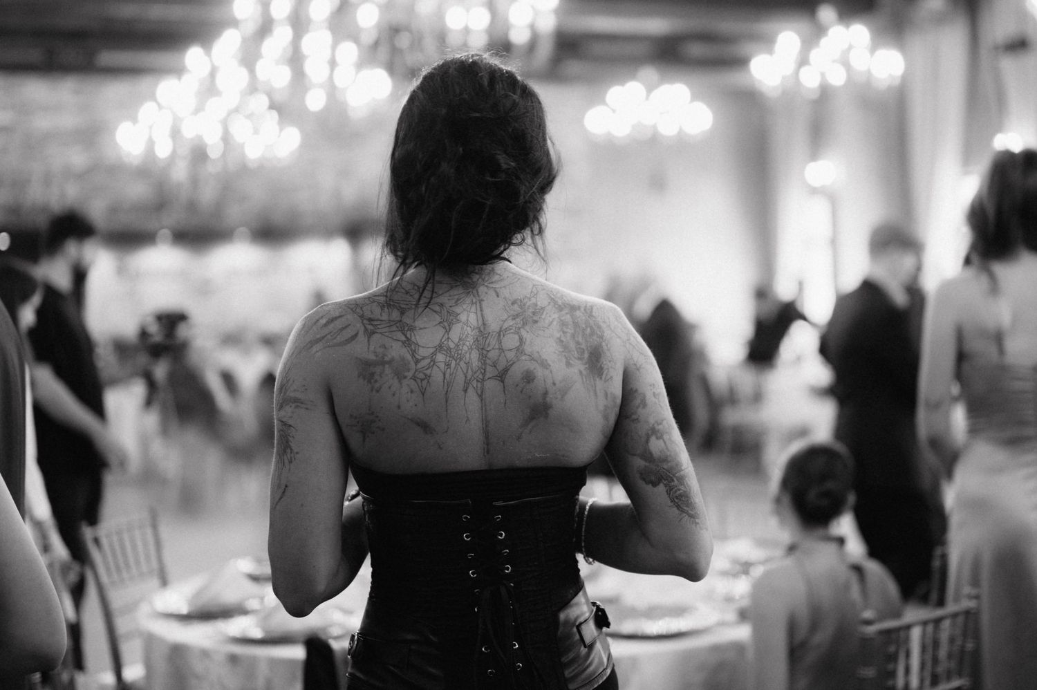 A person wearing a black corset with tattoos on their back stands in a dimly lit banquet hall with chandeliers.