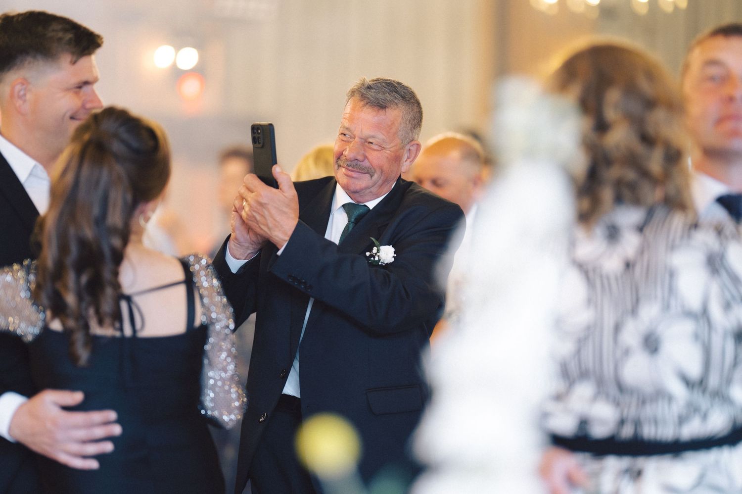 Wedding guests enjoying themselves at a formal reception as one person takes pictures with a mobile phone.