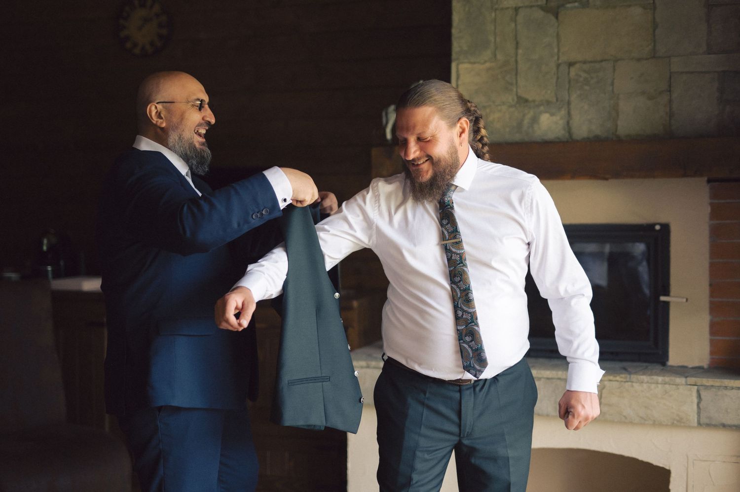 Two men stand in conversation, one in a navy suit and one in a white dress shirt and tie, sharing a lighthearted moment.