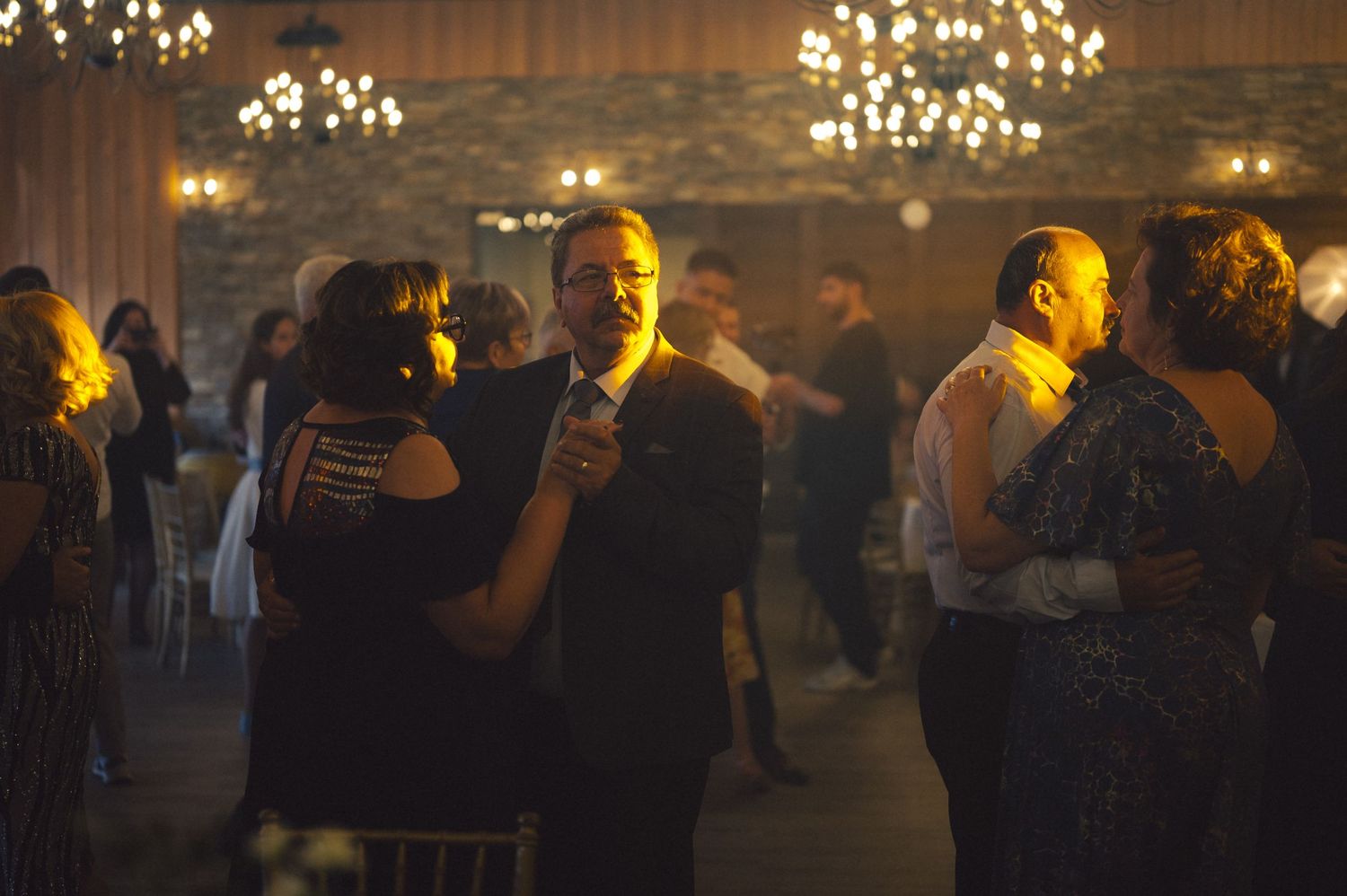 Couples slow dance together in a dimly lit ballroom with elegant chandeliers overhead creating a romantic atmosphere.